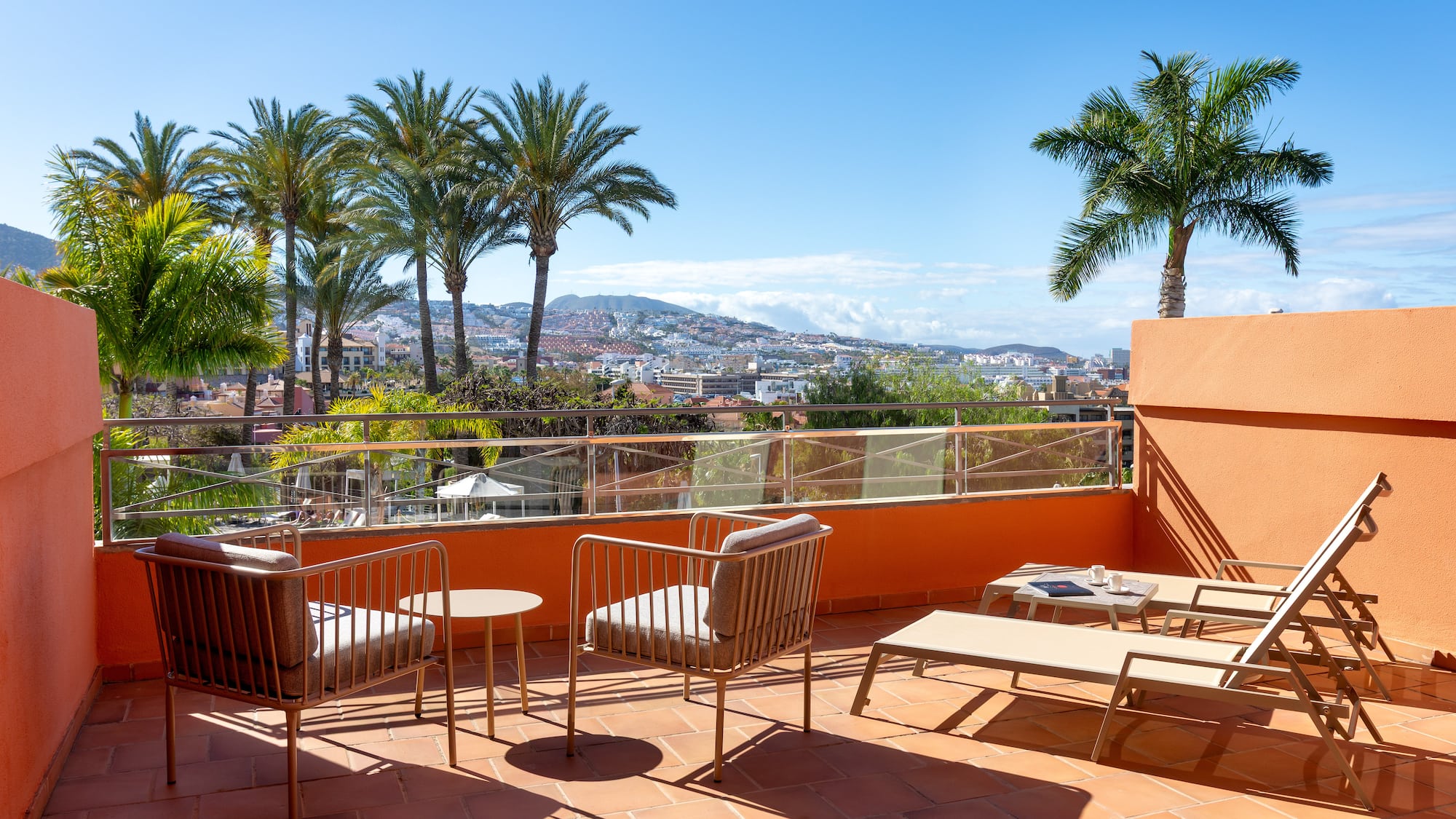 Sunny resort balcony with lounge chairs, palm trees, and city view.