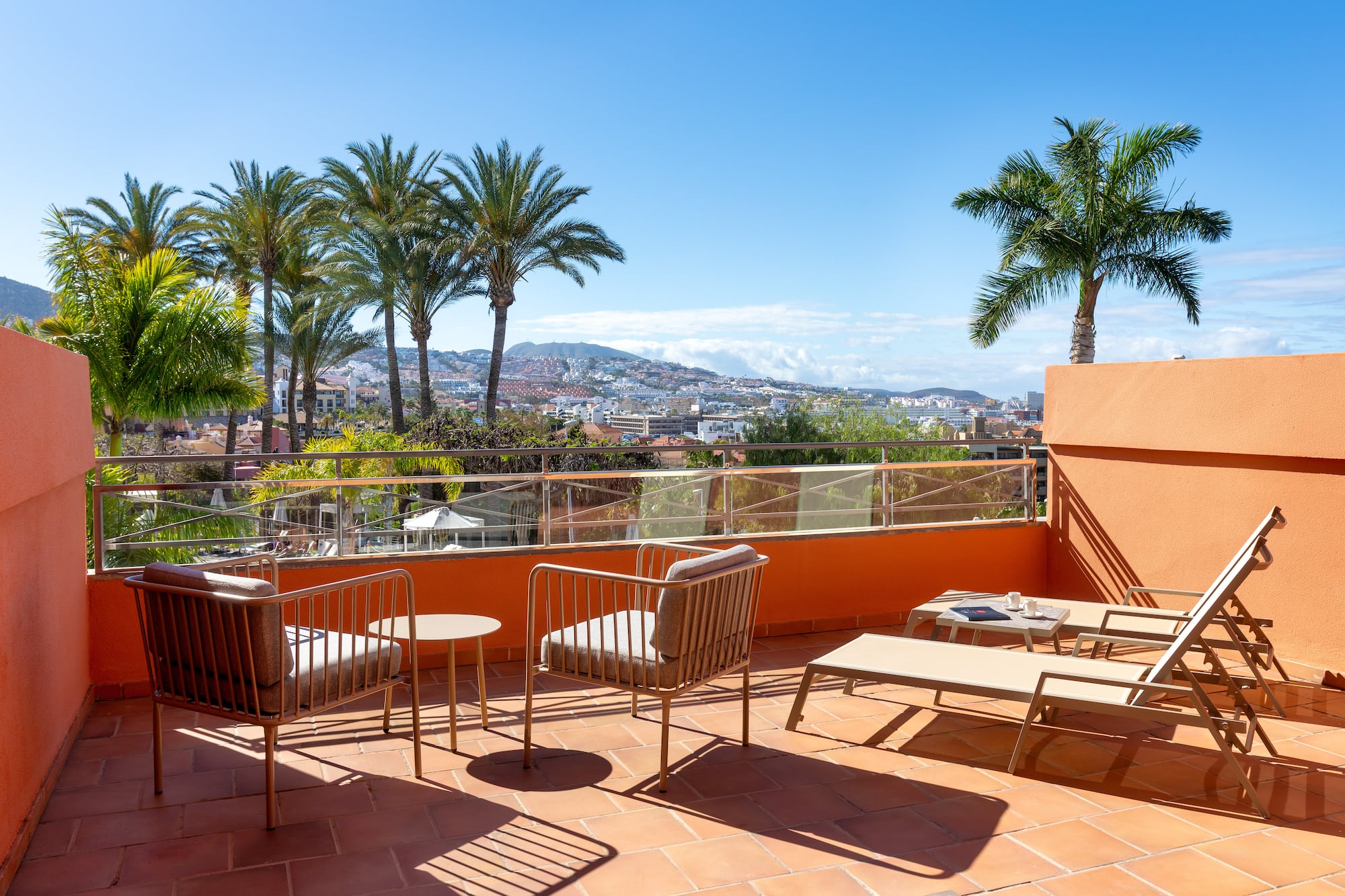 Sunny resort balcony with lounge chairs, palm trees, and city view.