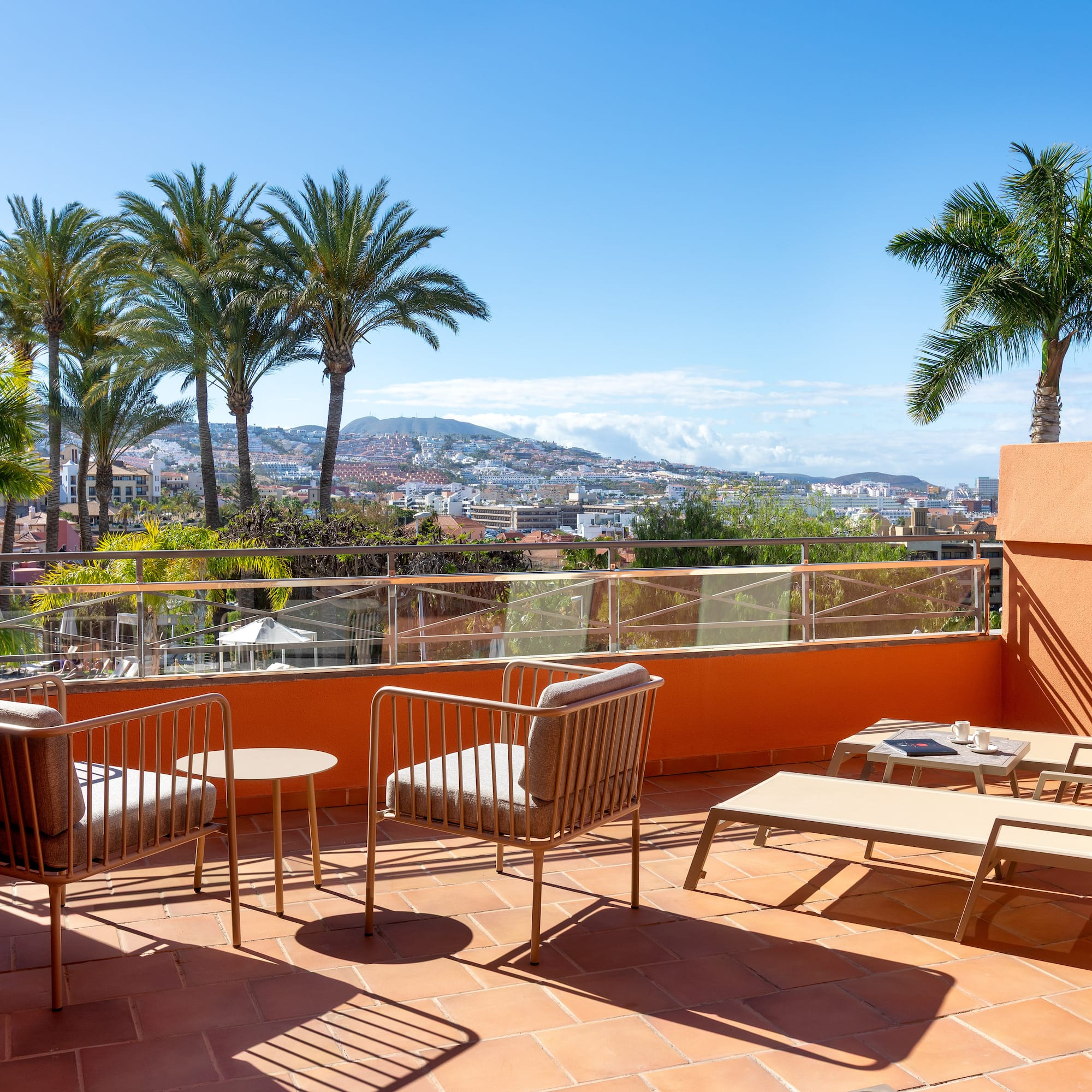 Sunny resort balcony with lounge chairs, palm trees, and city view.
