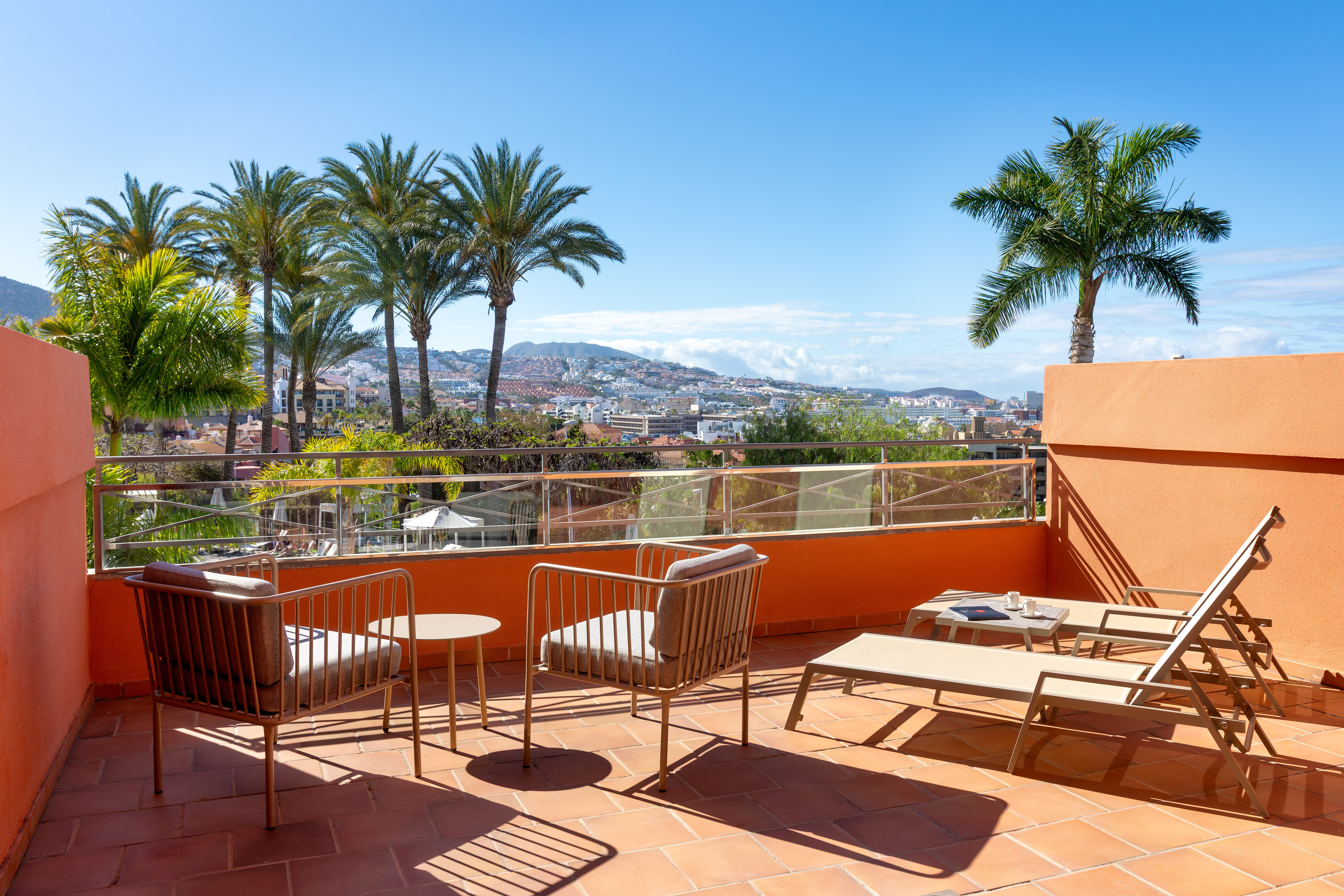 Sunny resort balcony with lounge chairs, palm trees, and city view.