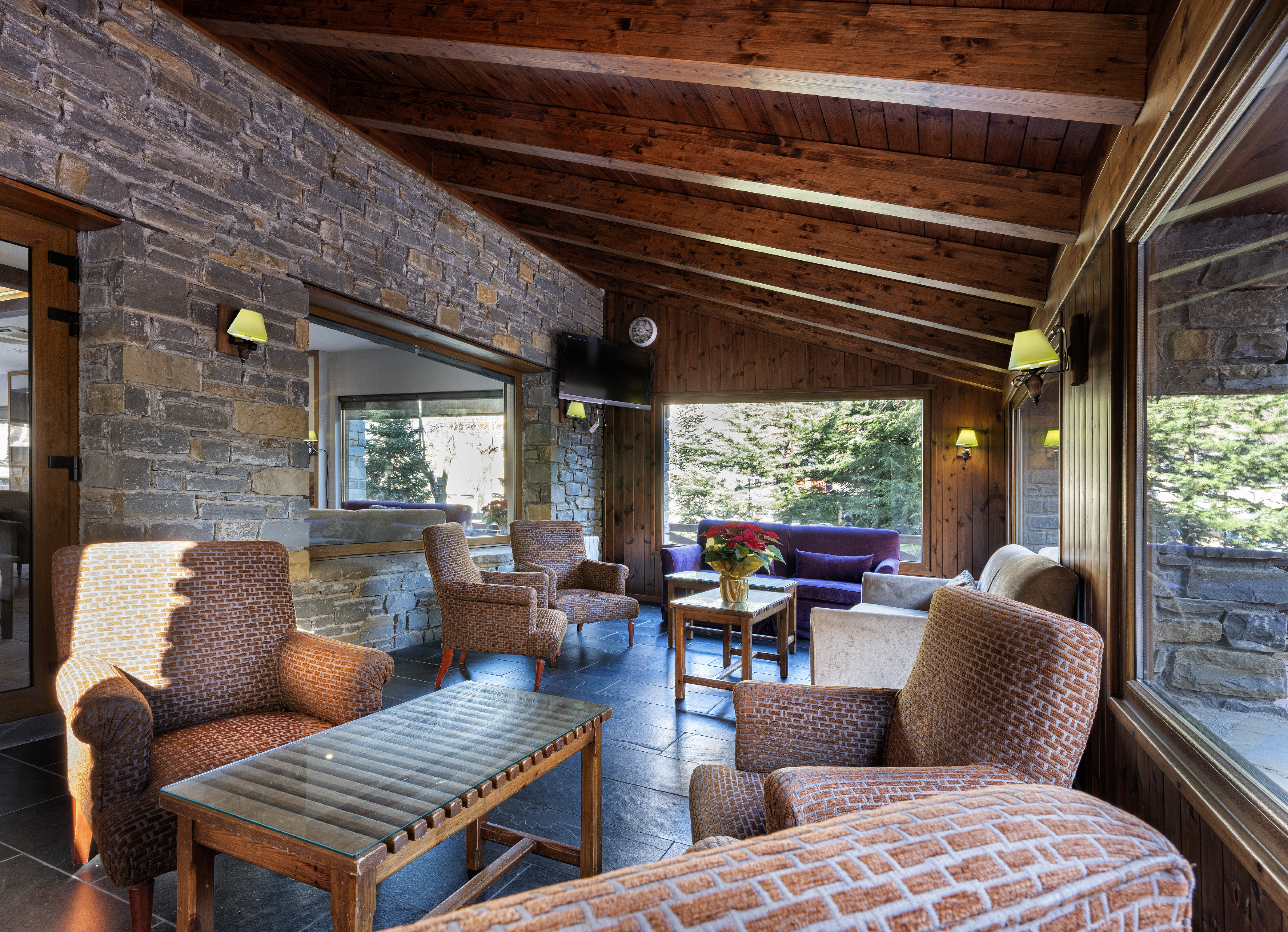 Rustic lodge living room with stone walls, wooden ceiling, armchairs, and forest views.