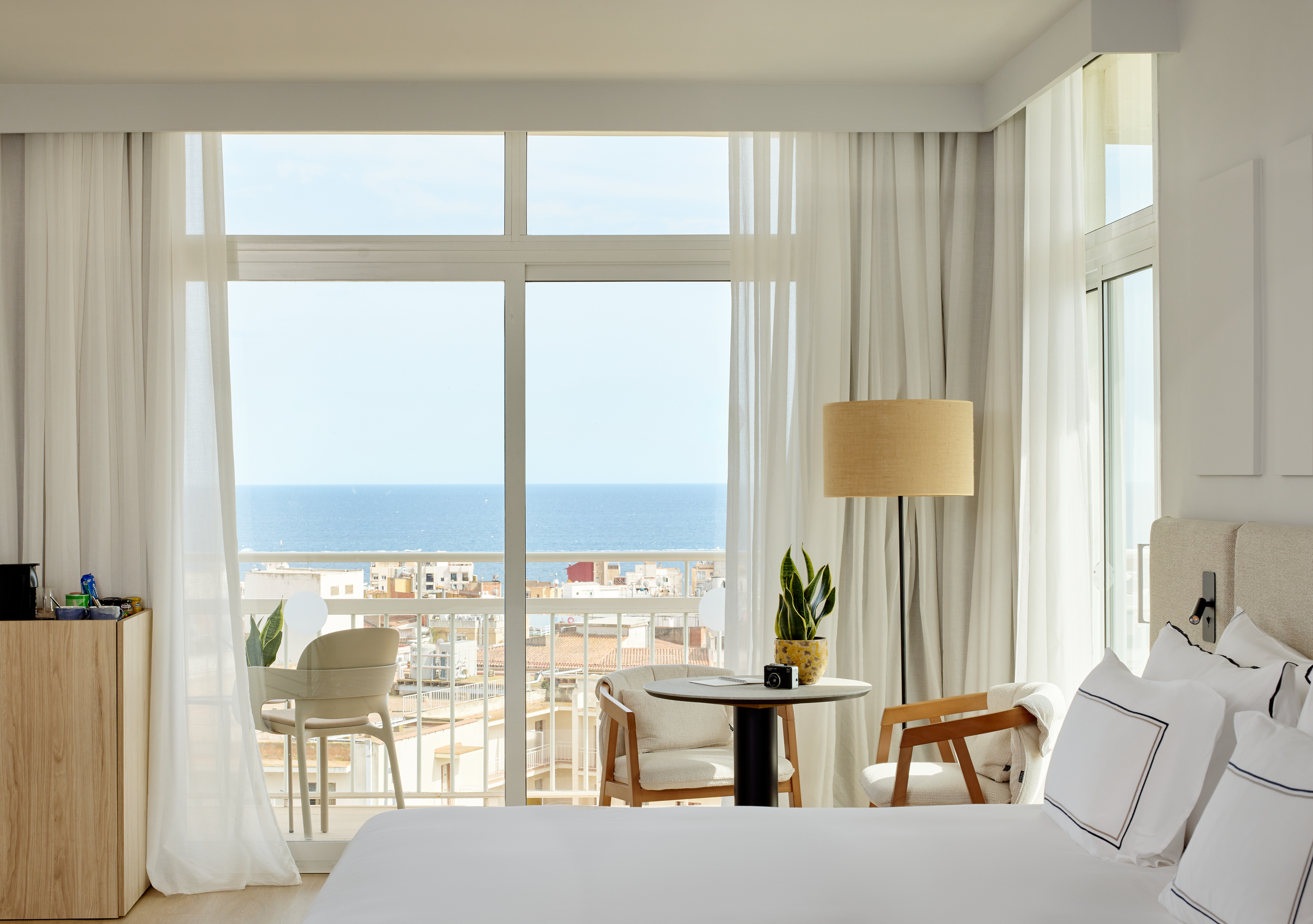 Modern hotel room with ocean view balcony, white decor, and city buildings.
