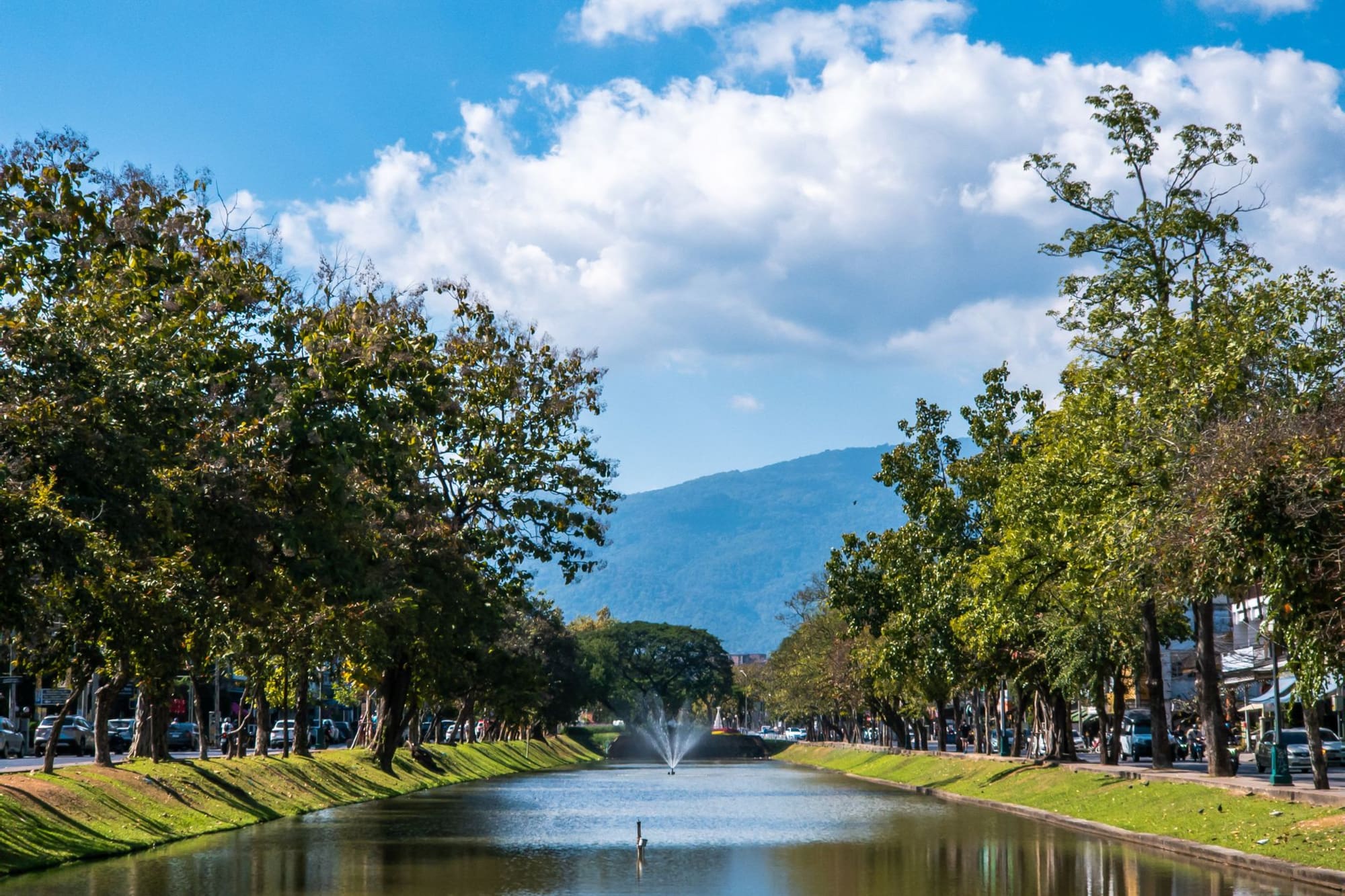Tree-lined canal with fountain and distant mountains under a blue sky.