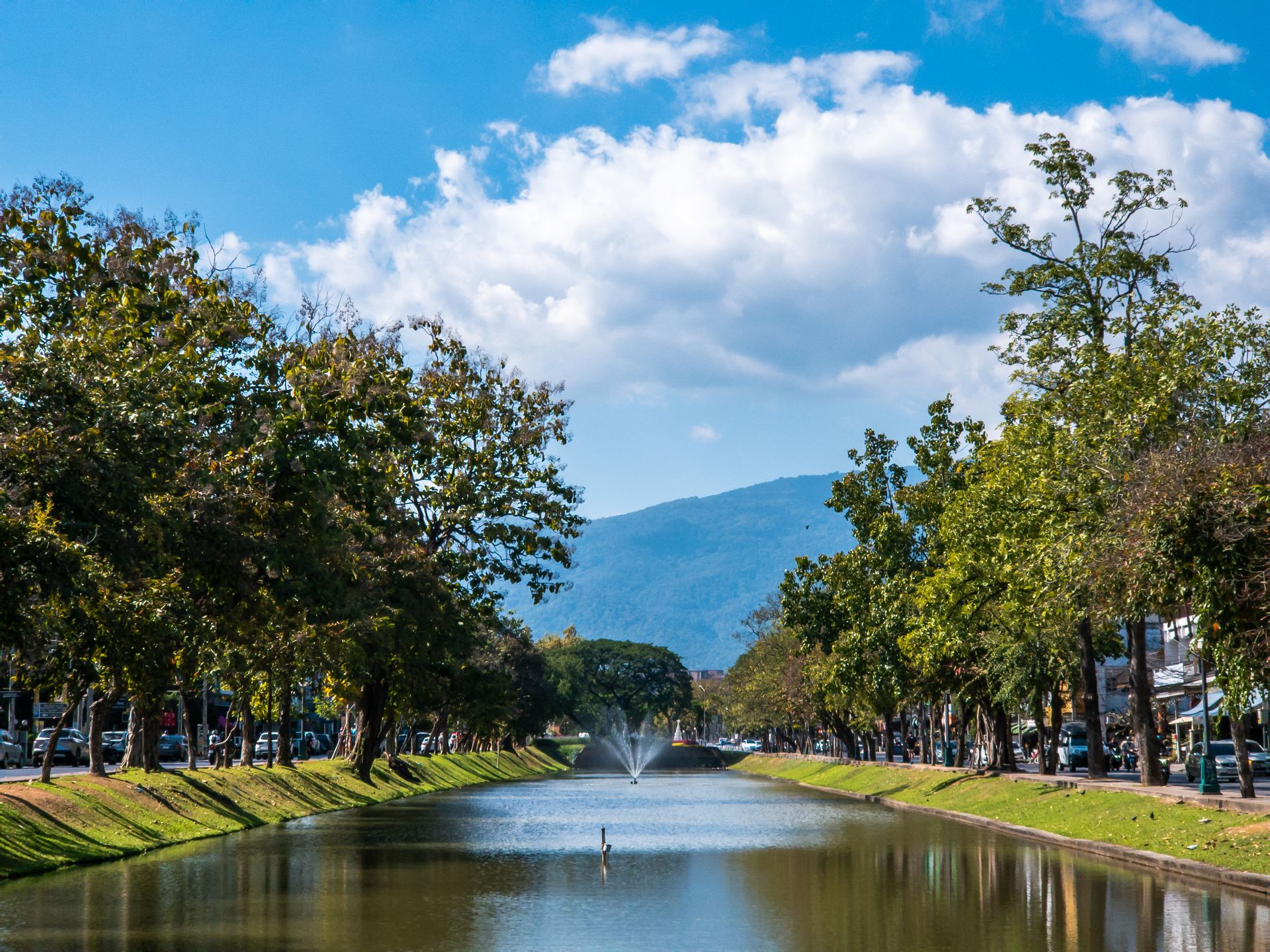 Tree-lined canal with fountain and distant mountains under a blue sky.