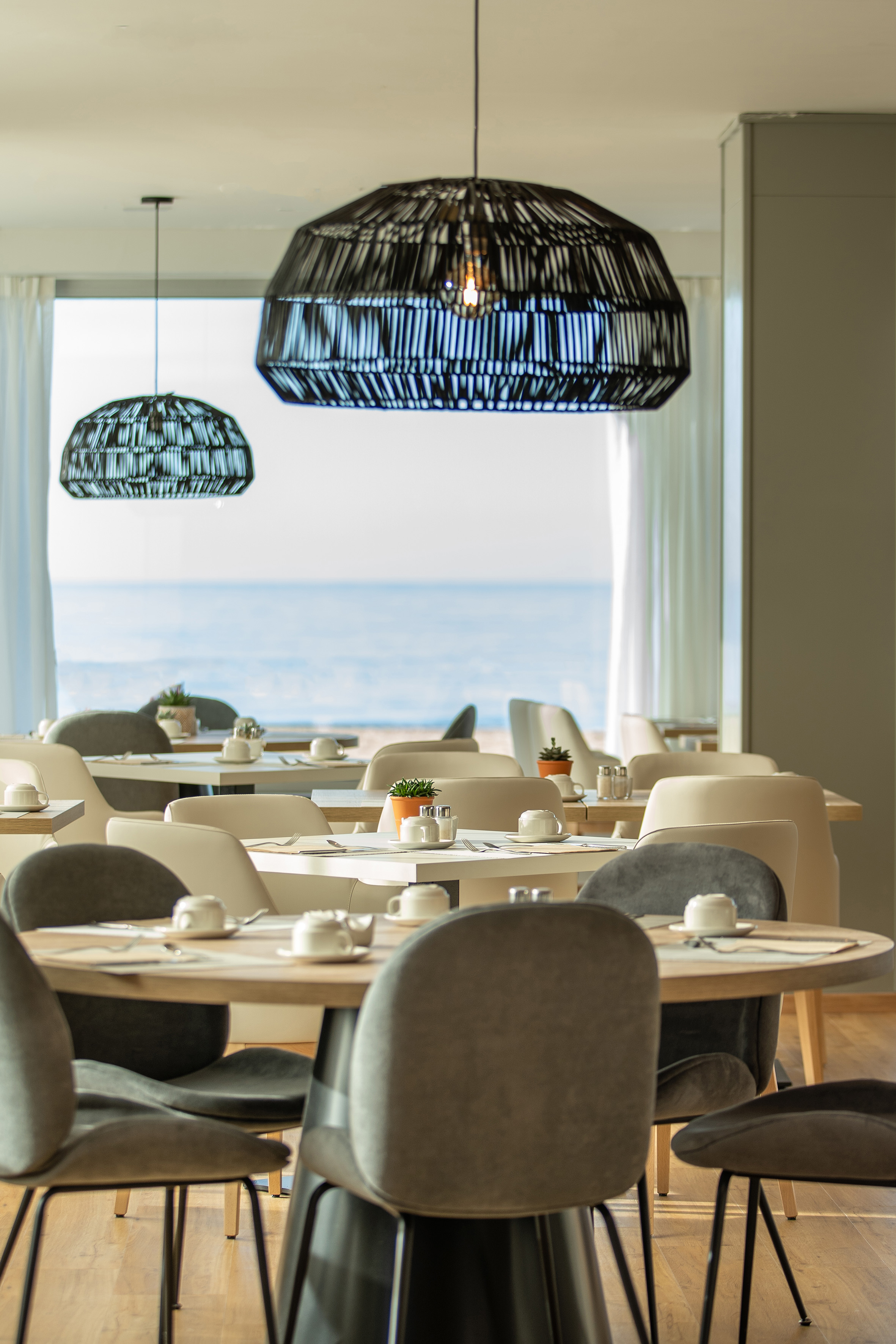 Modern dining area overlooking sea, set tables, chairs, woven pendant lights.