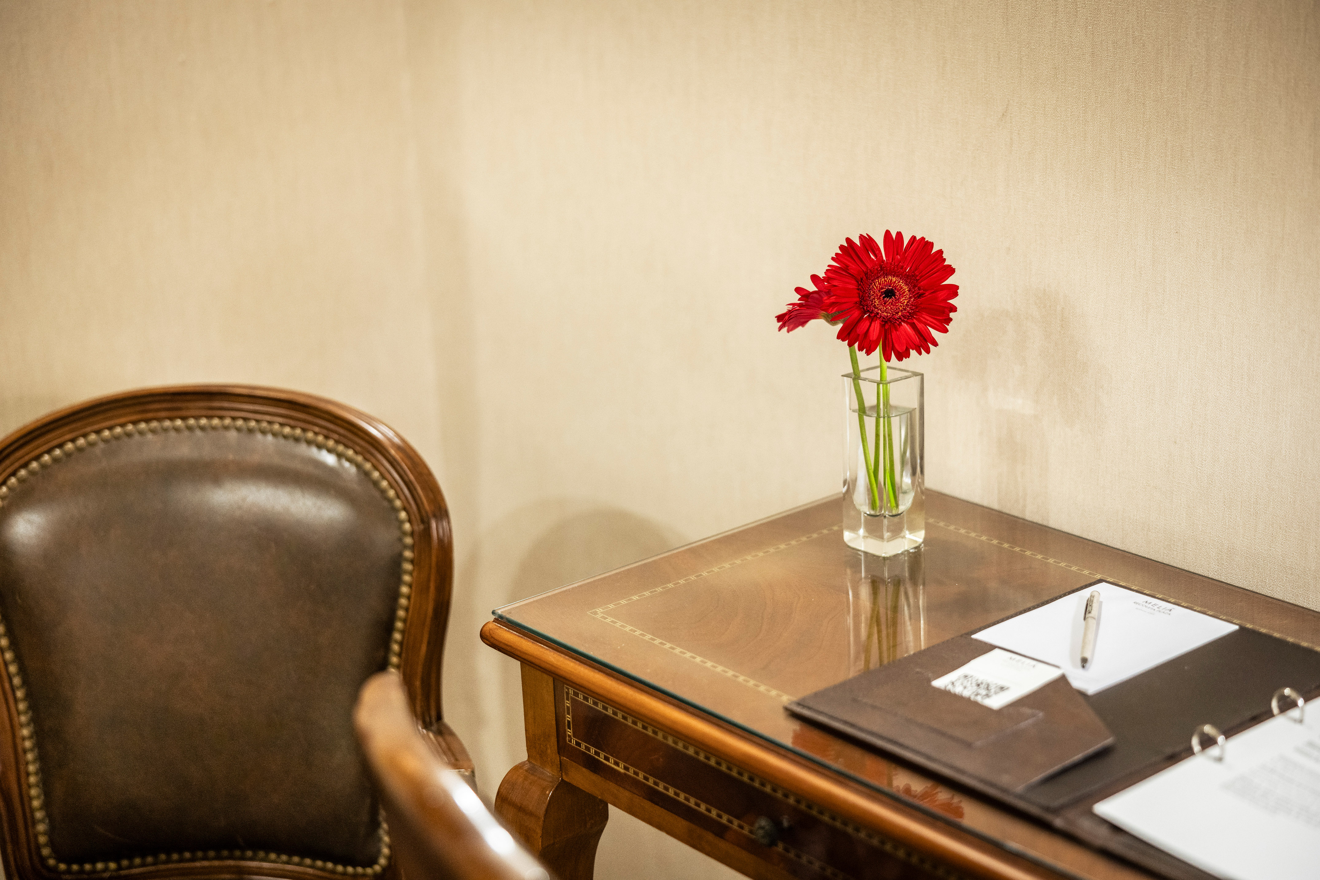 Red gerbera flowers on hotel desk with leather armchair and stationery.