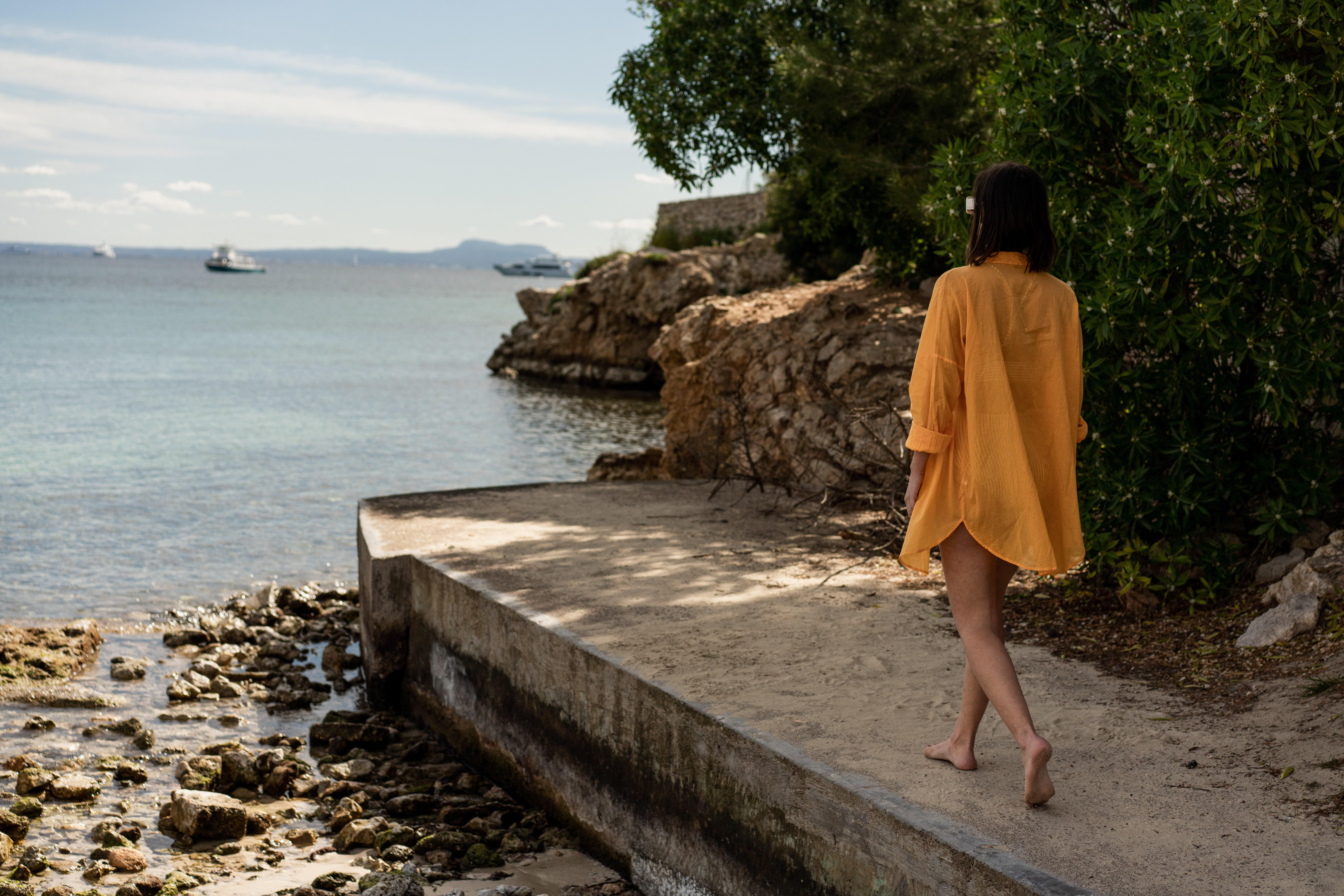 Woman in orange cover-up walking barefoot on a seaside path with boats.