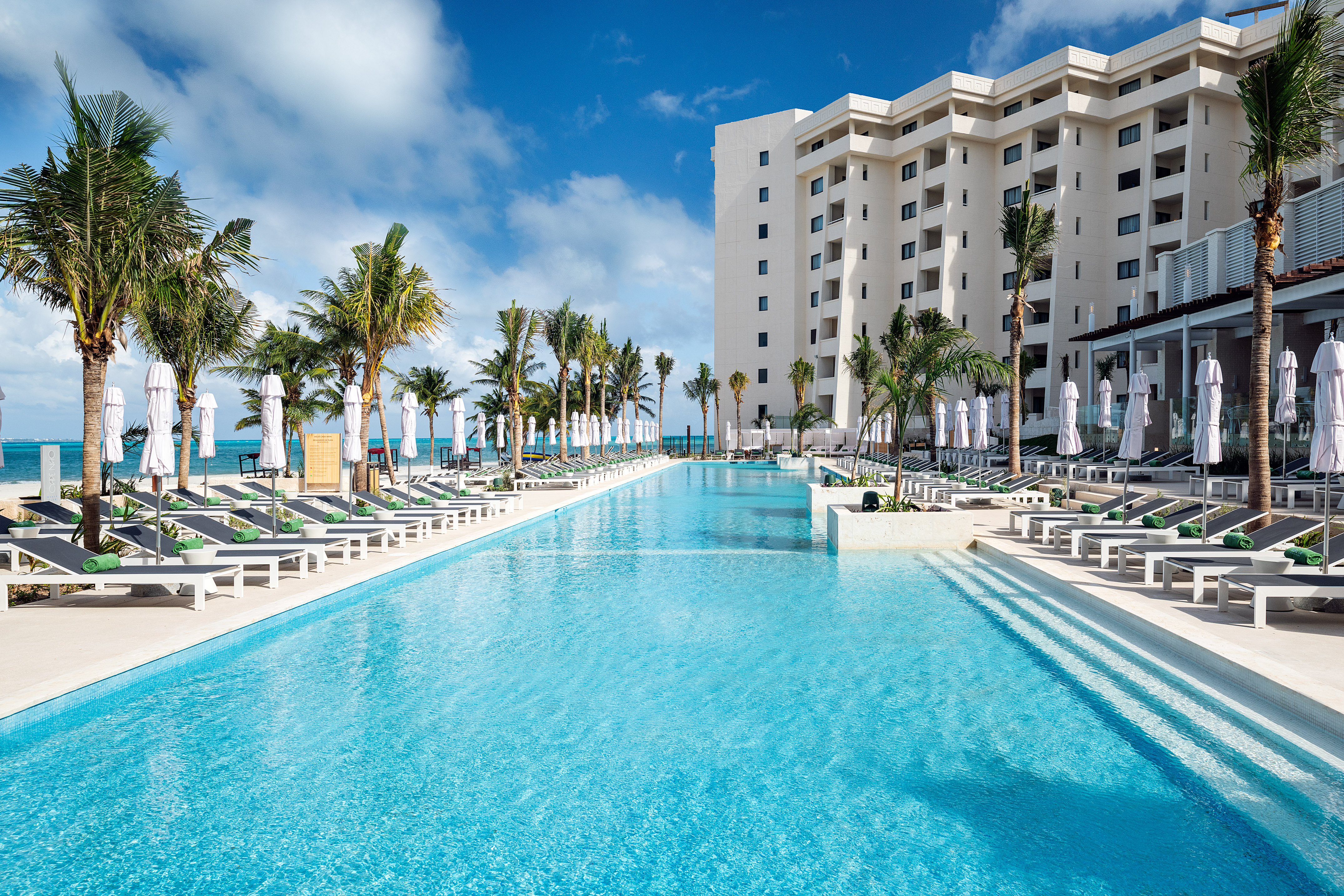 Tropical resort pool, palm trees, lounge chairs, and large hotel.