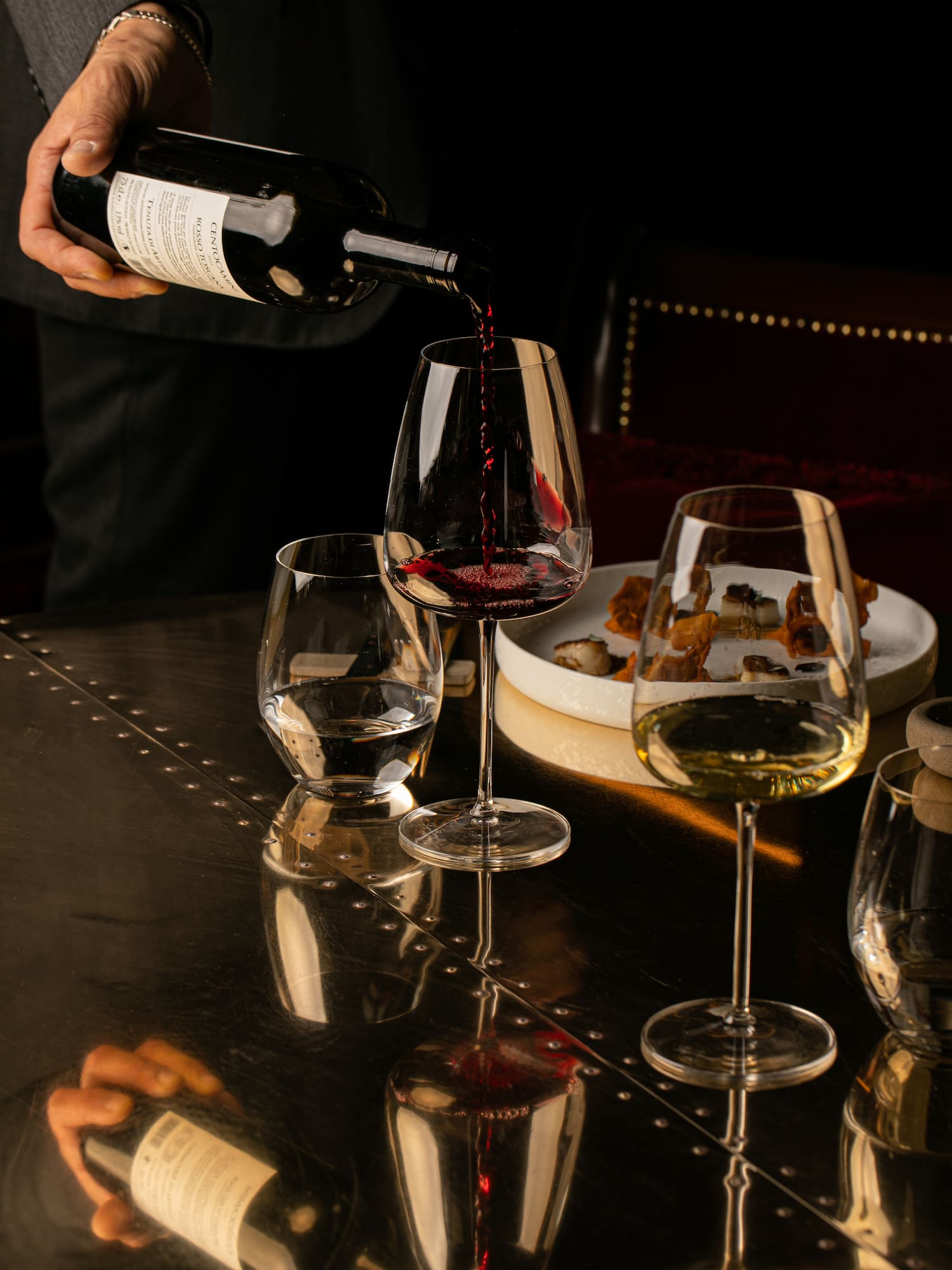 Sommelier pouring red wine into a glass, with white wine and appetizers.