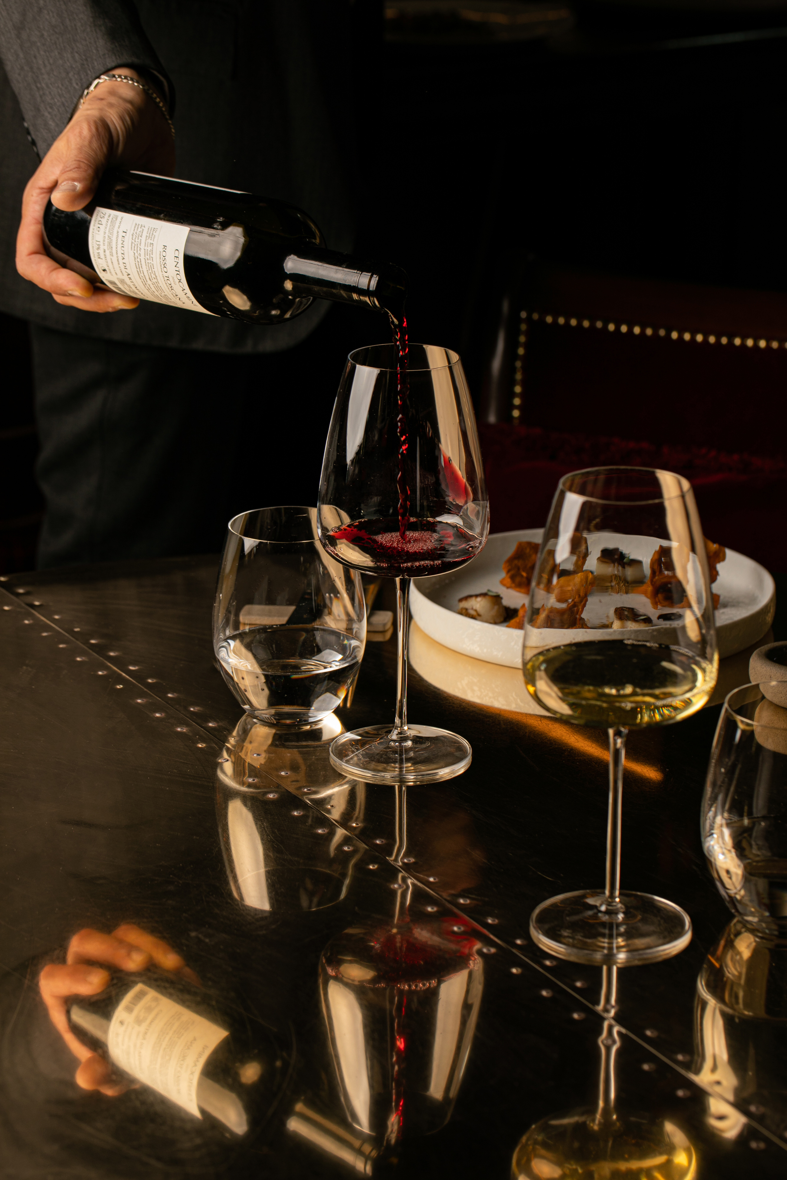 Sommelier pouring red wine into a glass, with white wine and appetizers.