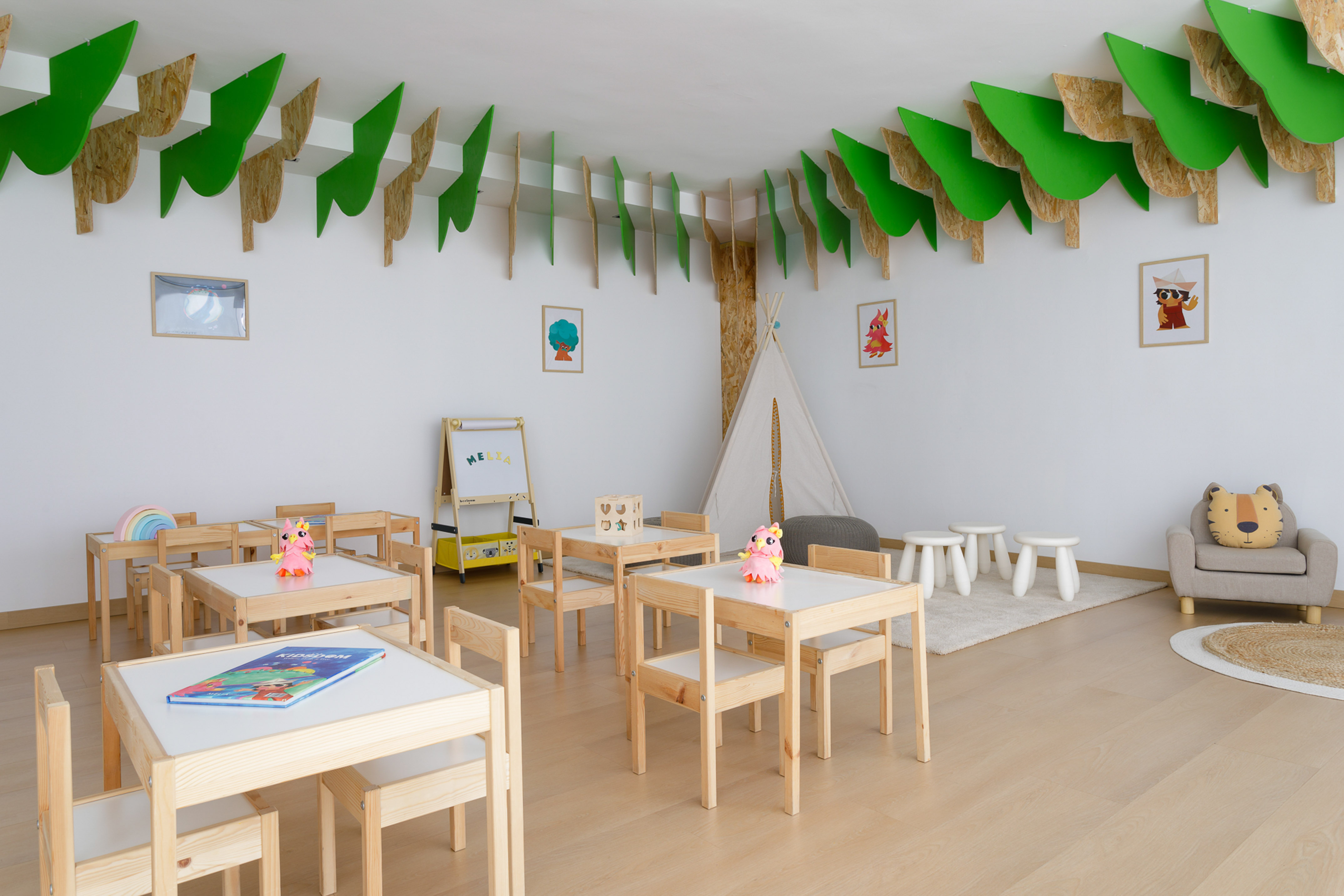Kids' classroom with wooden tables, chairs, teepee, and green leaf ceiling decor.