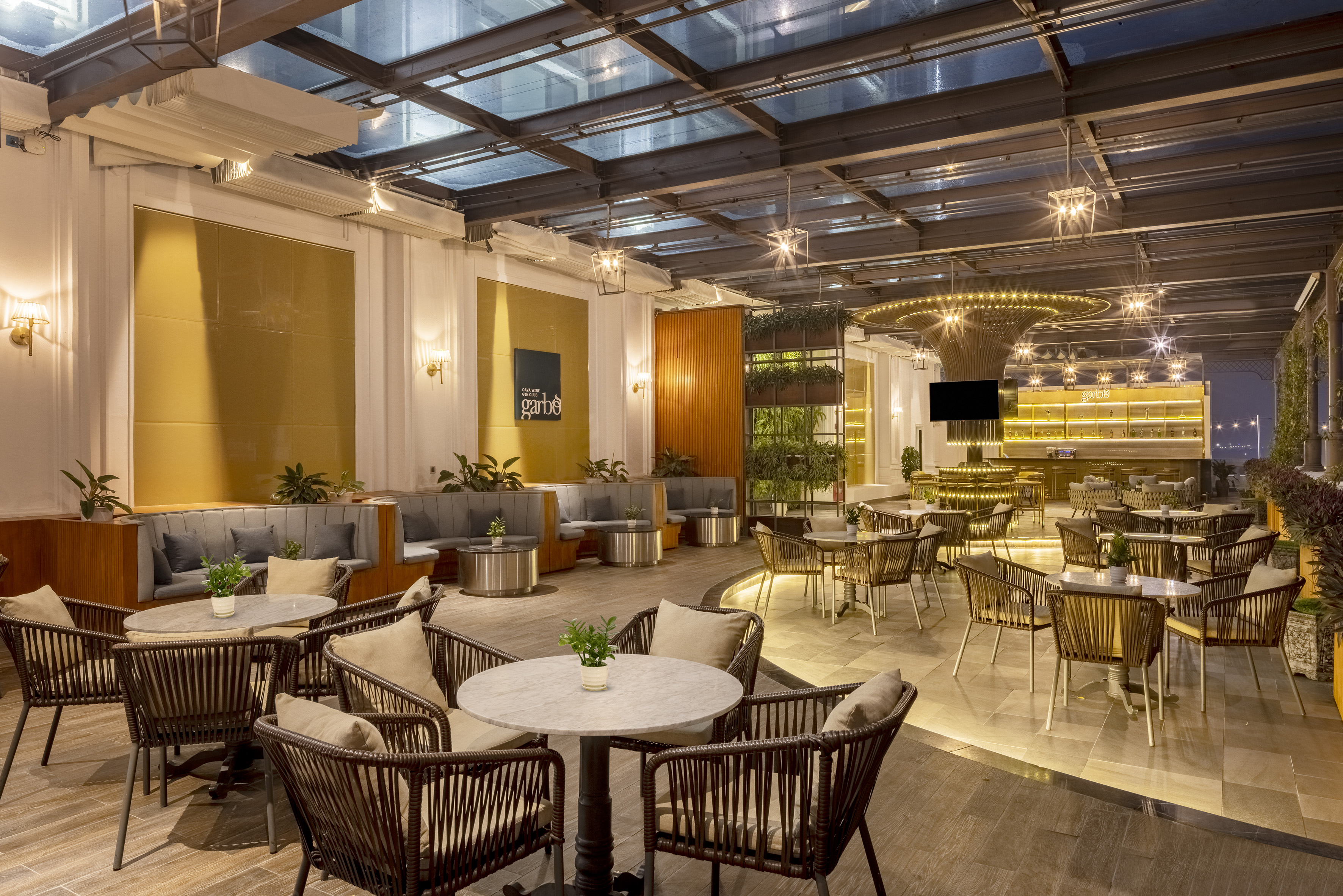 Modern rooftop restaurant with glass roof, rattan chairs, and illuminated bar.