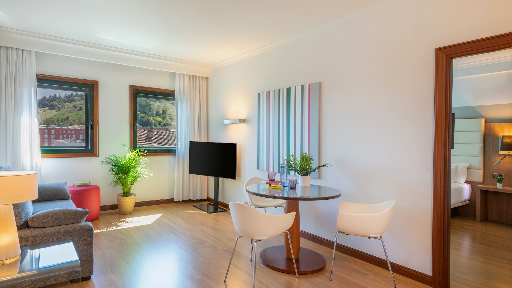 Modern hotel suite living room with sofa, TV, dining area, and bright windows.