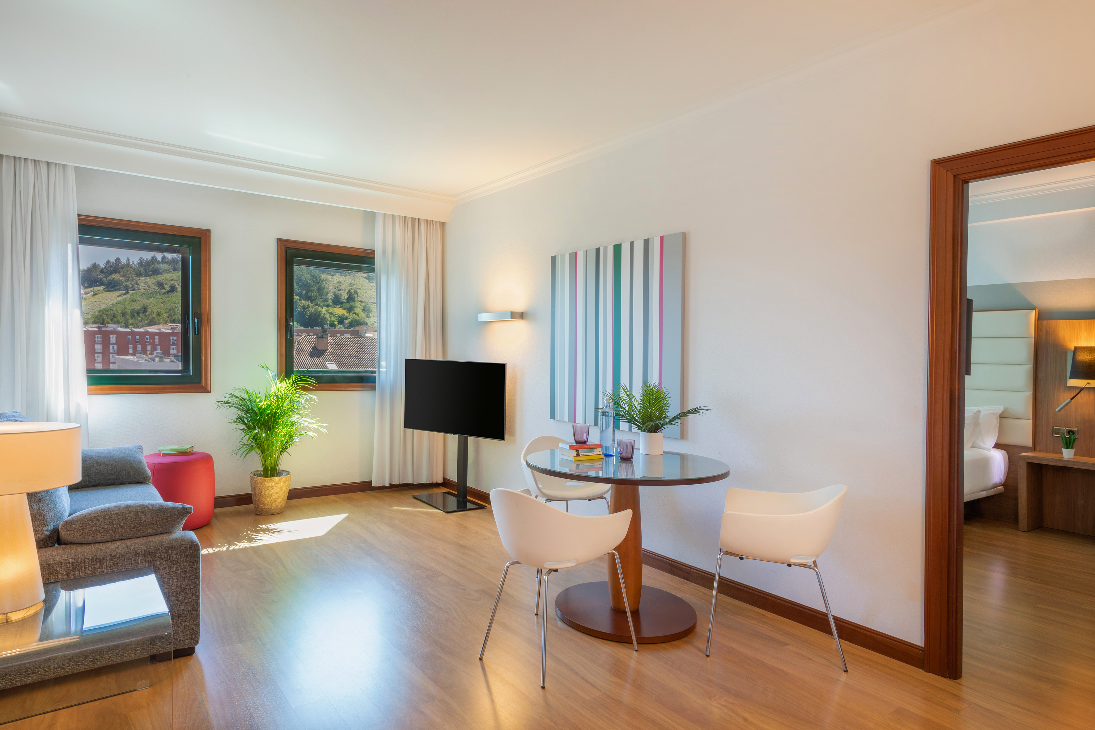 Modern hotel suite living room with sofa, TV, dining area, and bright windows.