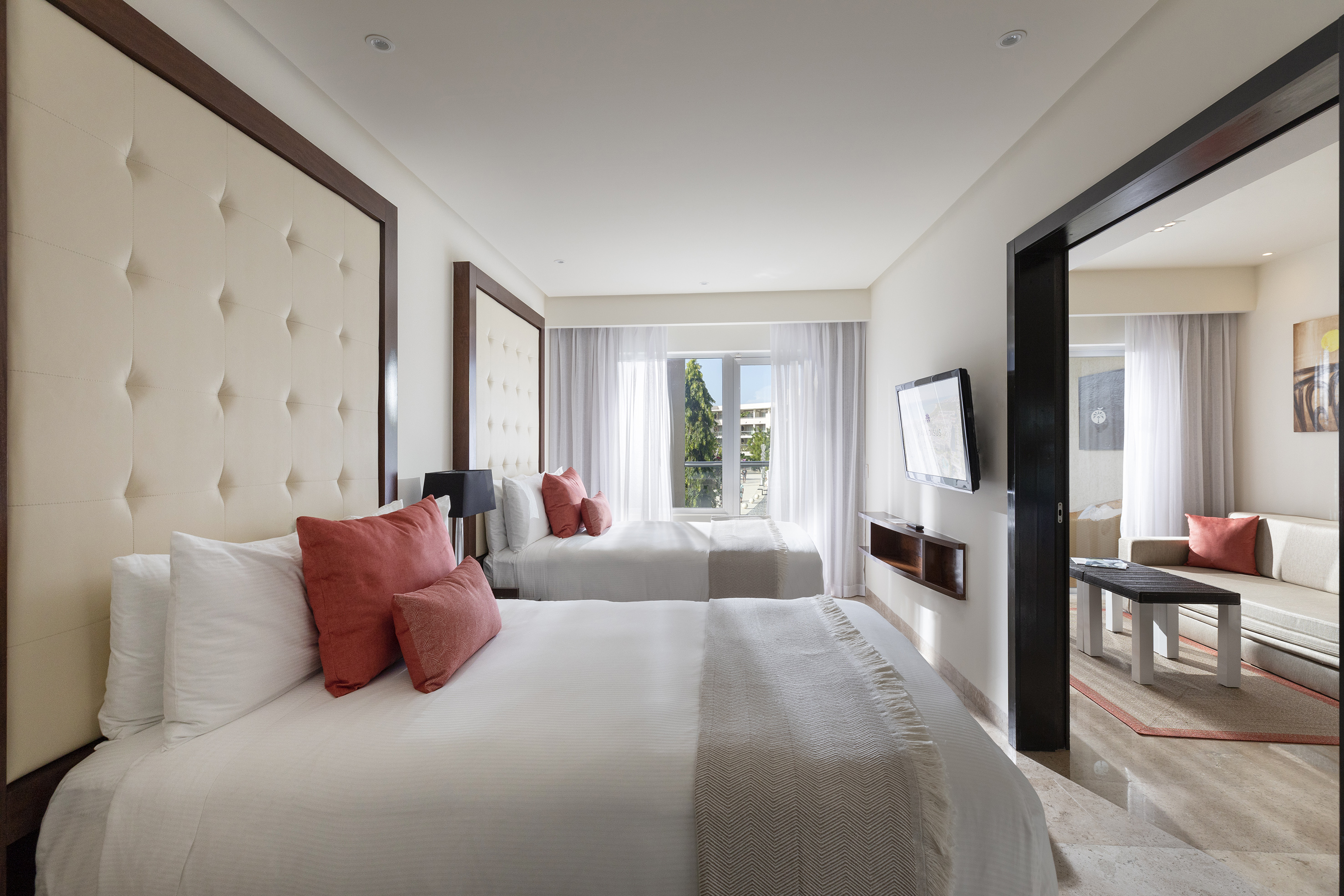 Modern hotel suite with two beds, coral pillows, tufted headboards, and living space.