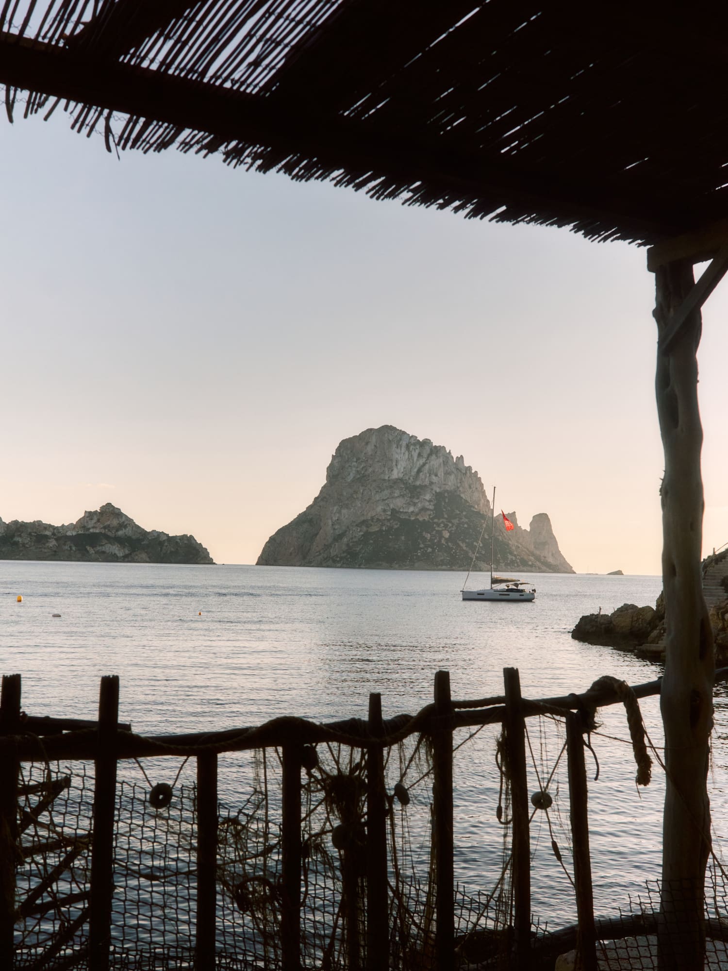 Es Vedrà island and sailboat at sunset, framed by a rustic beach bar, Ibiza.
