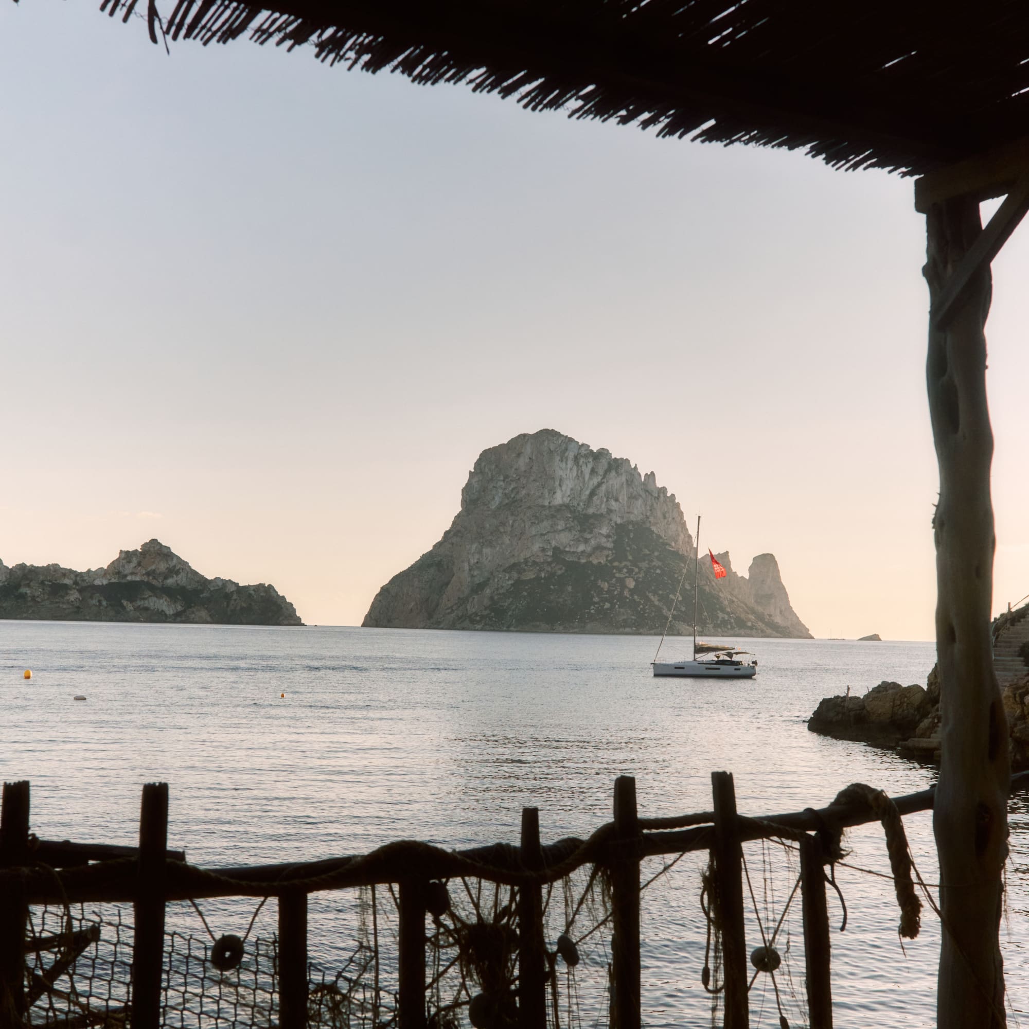 Es Vedrà island and sailboat at sunset, framed by a rustic beach bar, Ibiza.