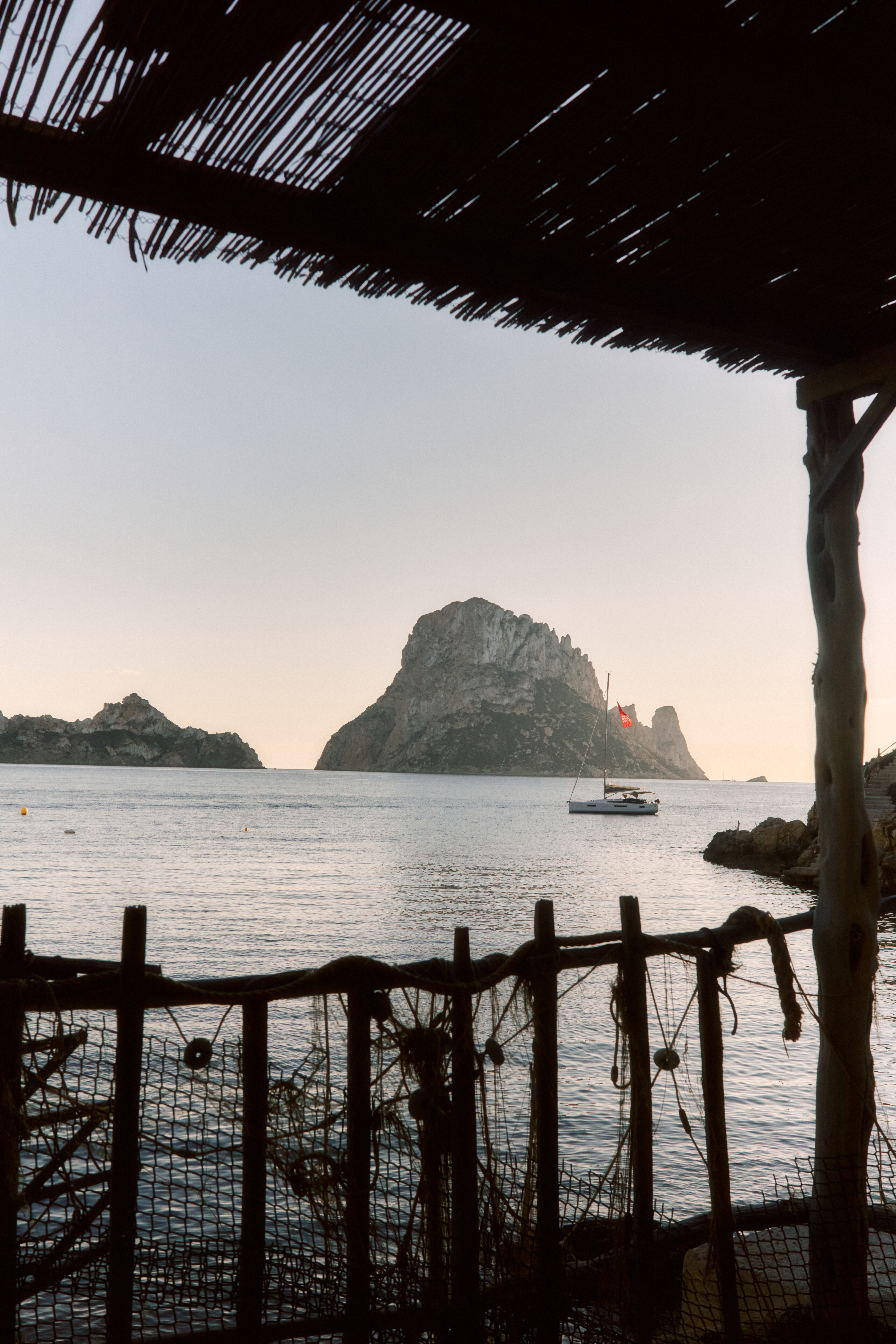 Es Vedrà island and sailboat at sunset, framed by a rustic beach bar, Ibiza.