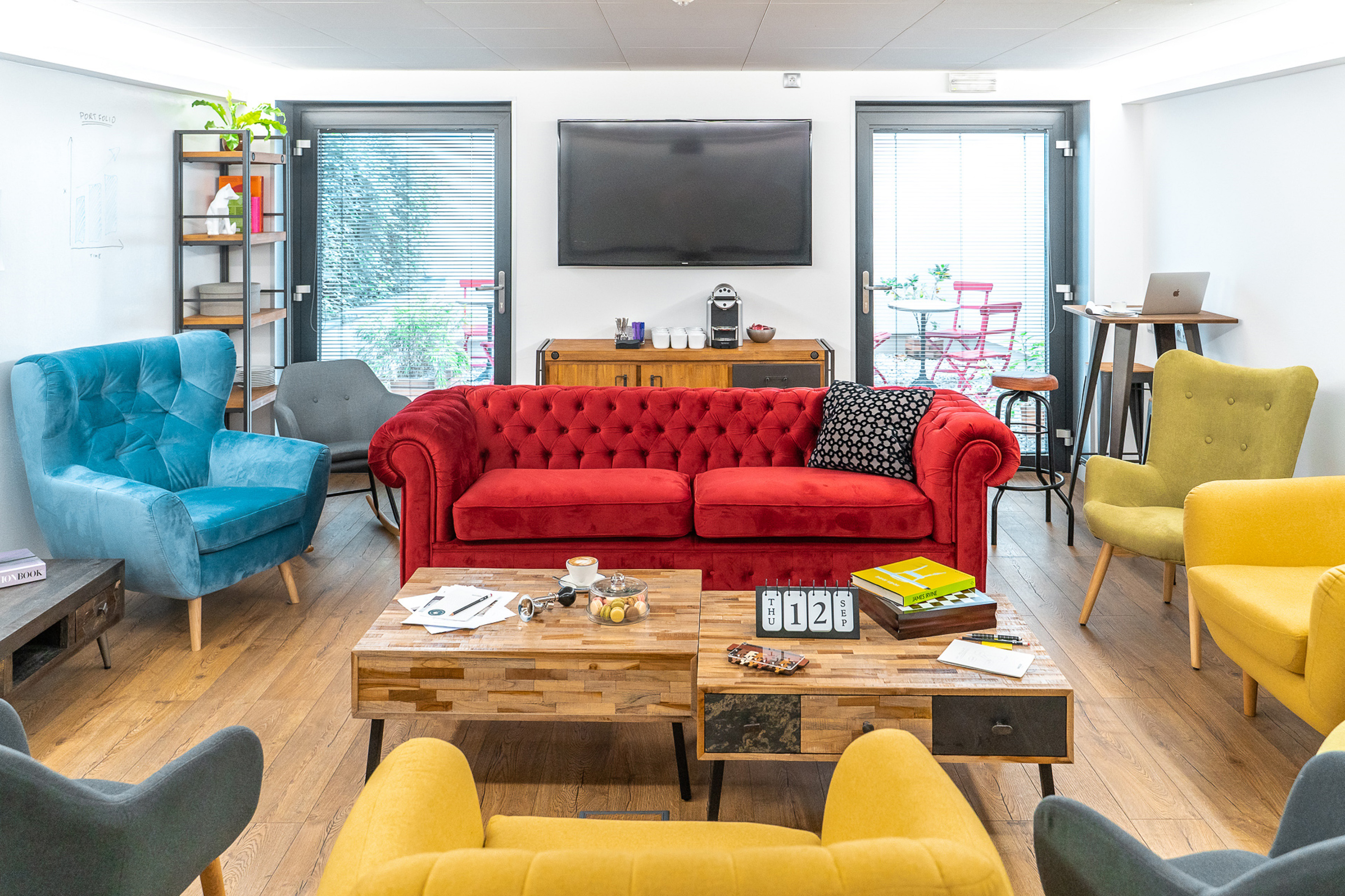 Modern office lounge with red sofa, blue, yellow chairs, coffee tables, and TV.