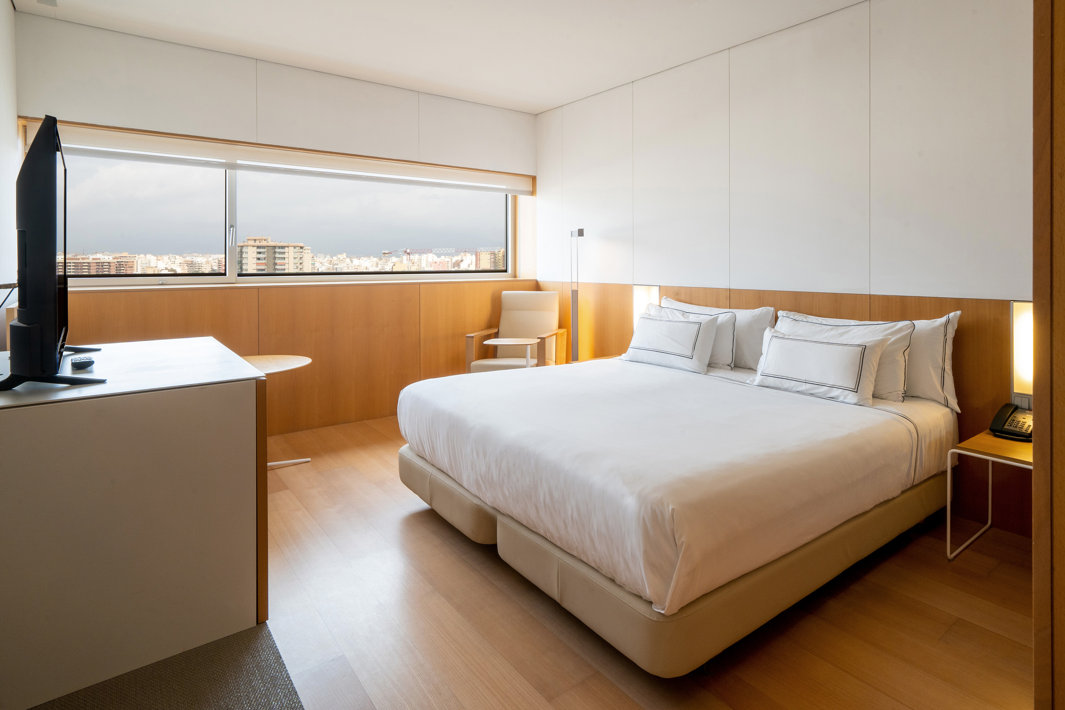 Modern hotel bedroom with large bed, city view window, light wood, and TV.