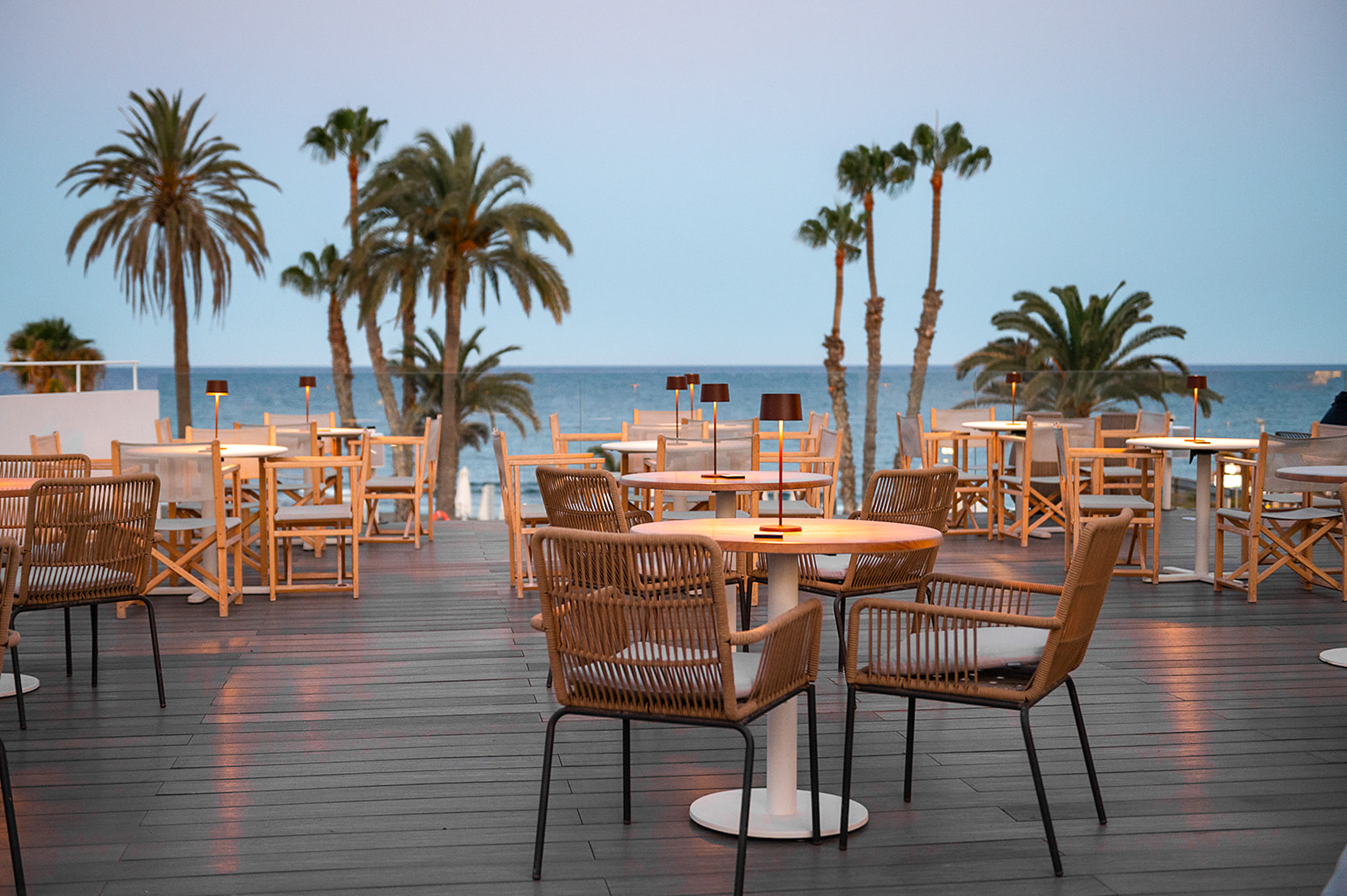 Outdoor restaurant patio with tables, chairs, and palm trees by the sea at dusk.