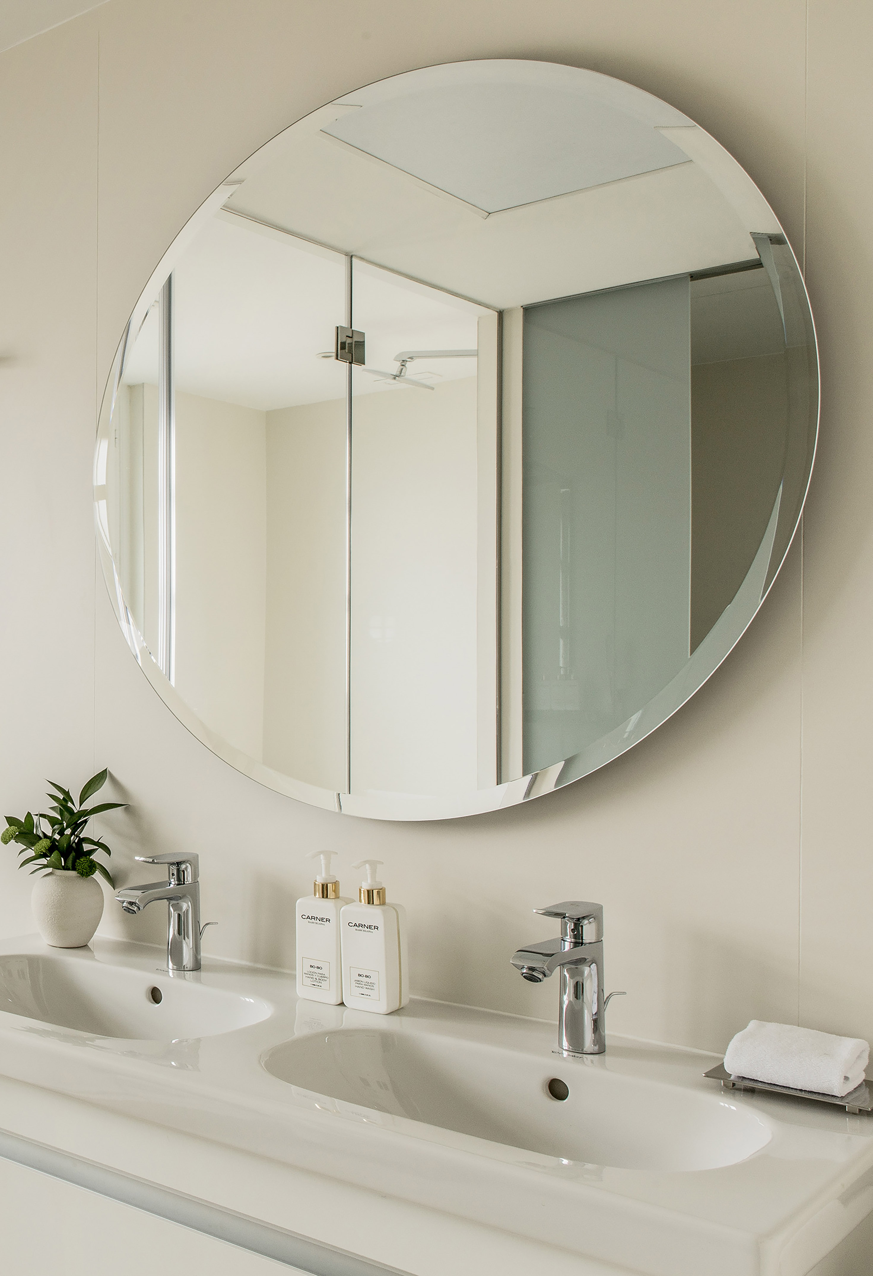 Round bathroom mirror above a double vanity with sinks and chrome faucets.