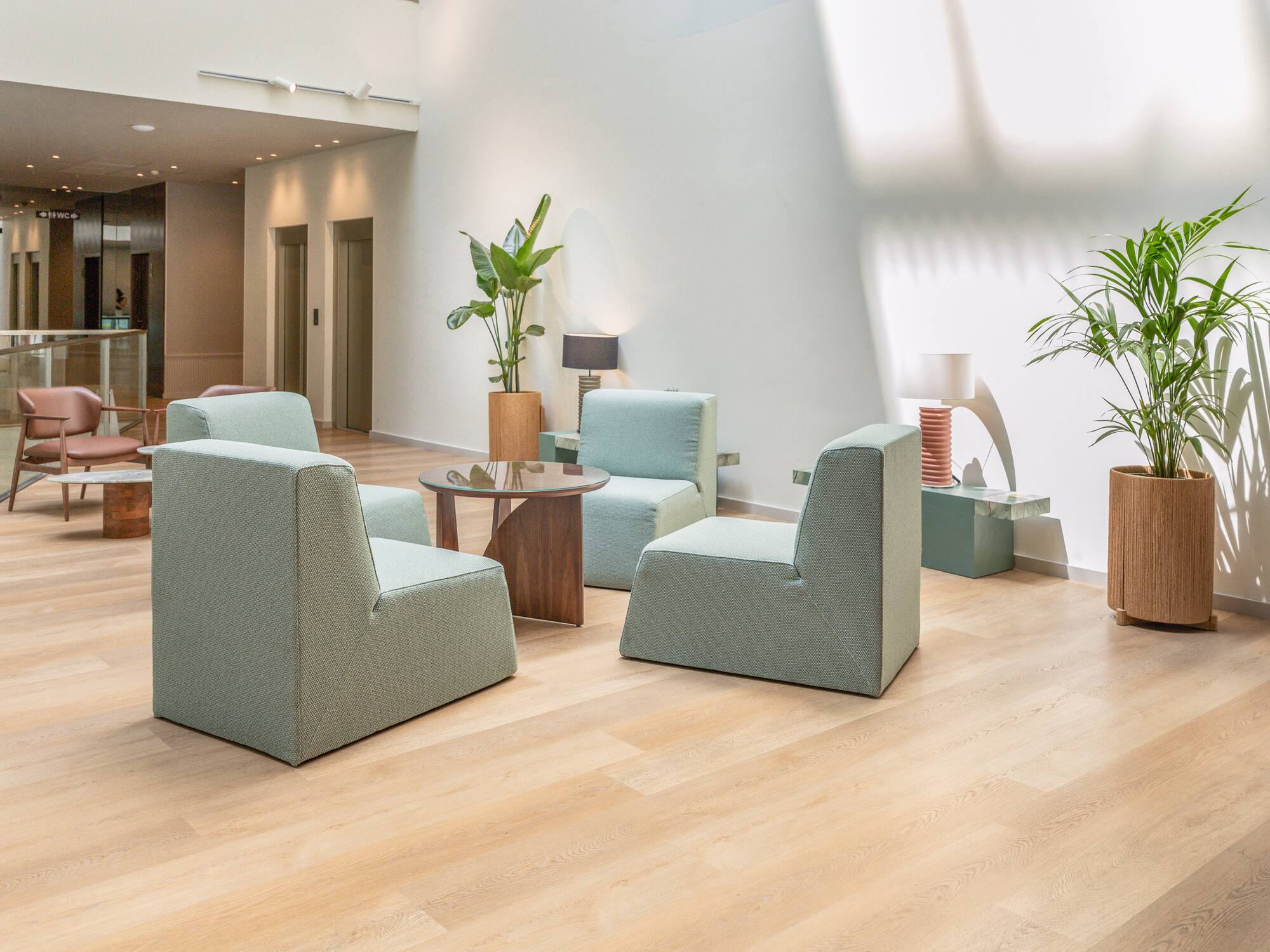 Modern lobby with light green armchairs, wooden floor, and potted plants.