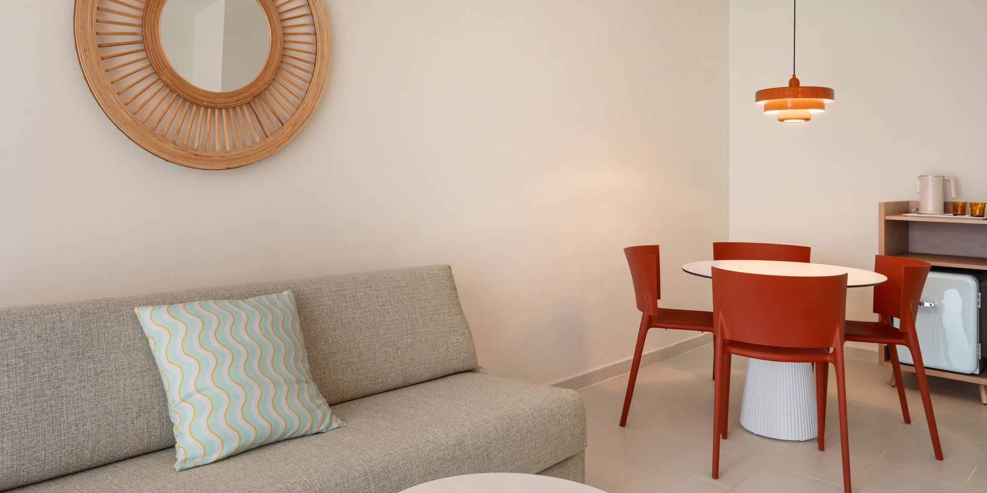 Hotel suite with modern grey sofa, red dining set, and rattan mirror.
