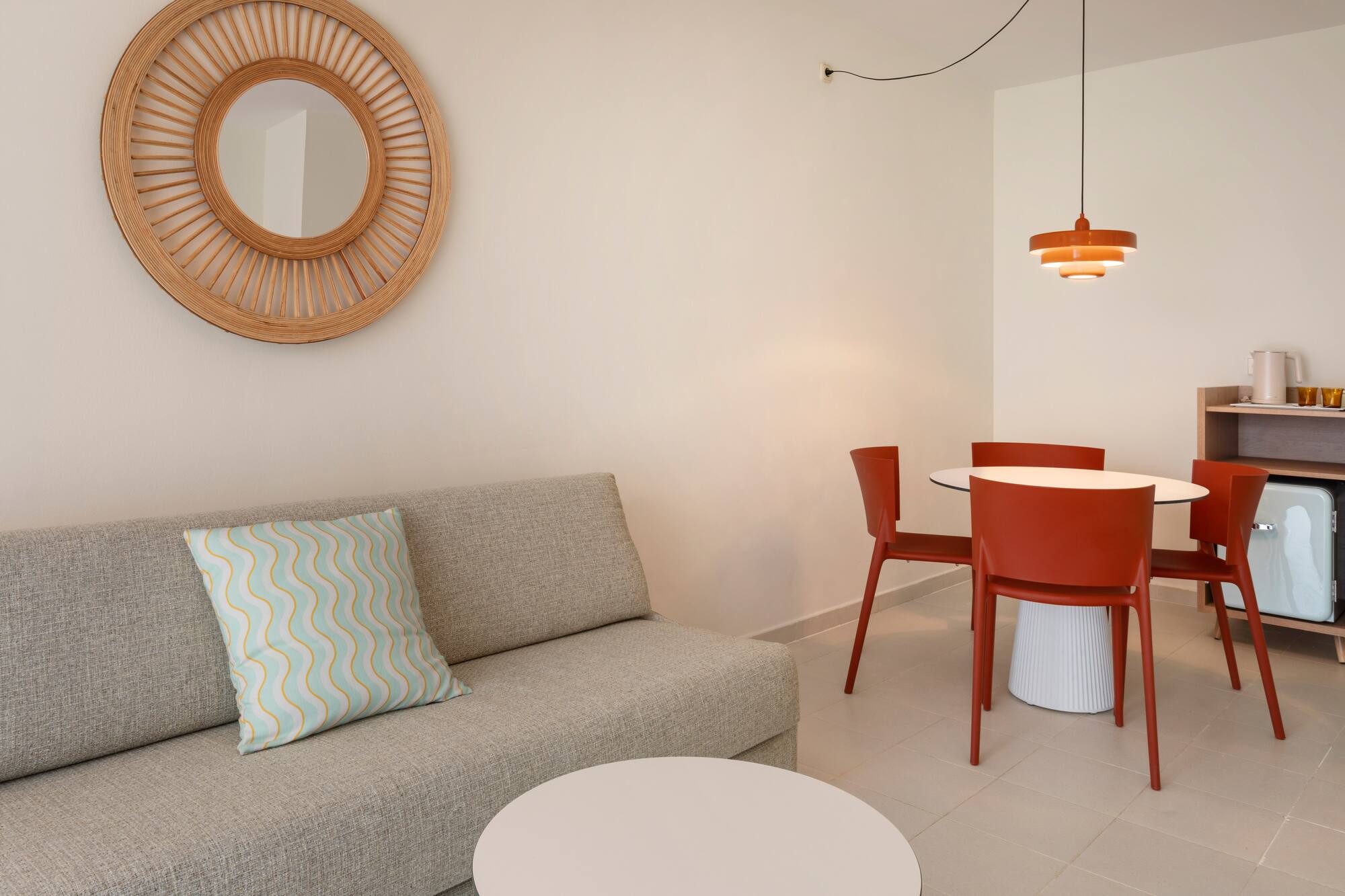 Hotel suite with modern grey sofa, red dining set, and rattan mirror.