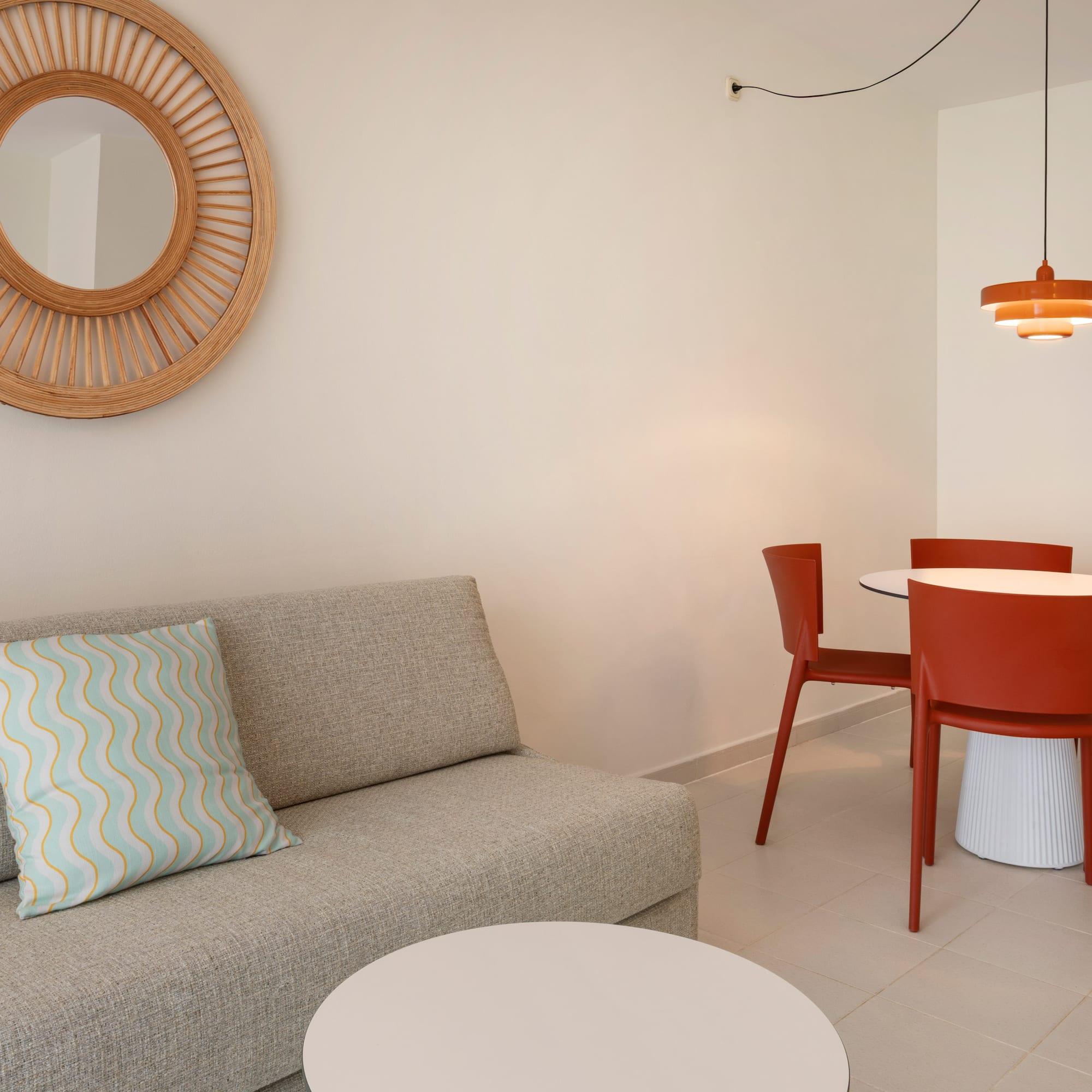 Hotel suite with modern grey sofa, red dining set, and rattan mirror.