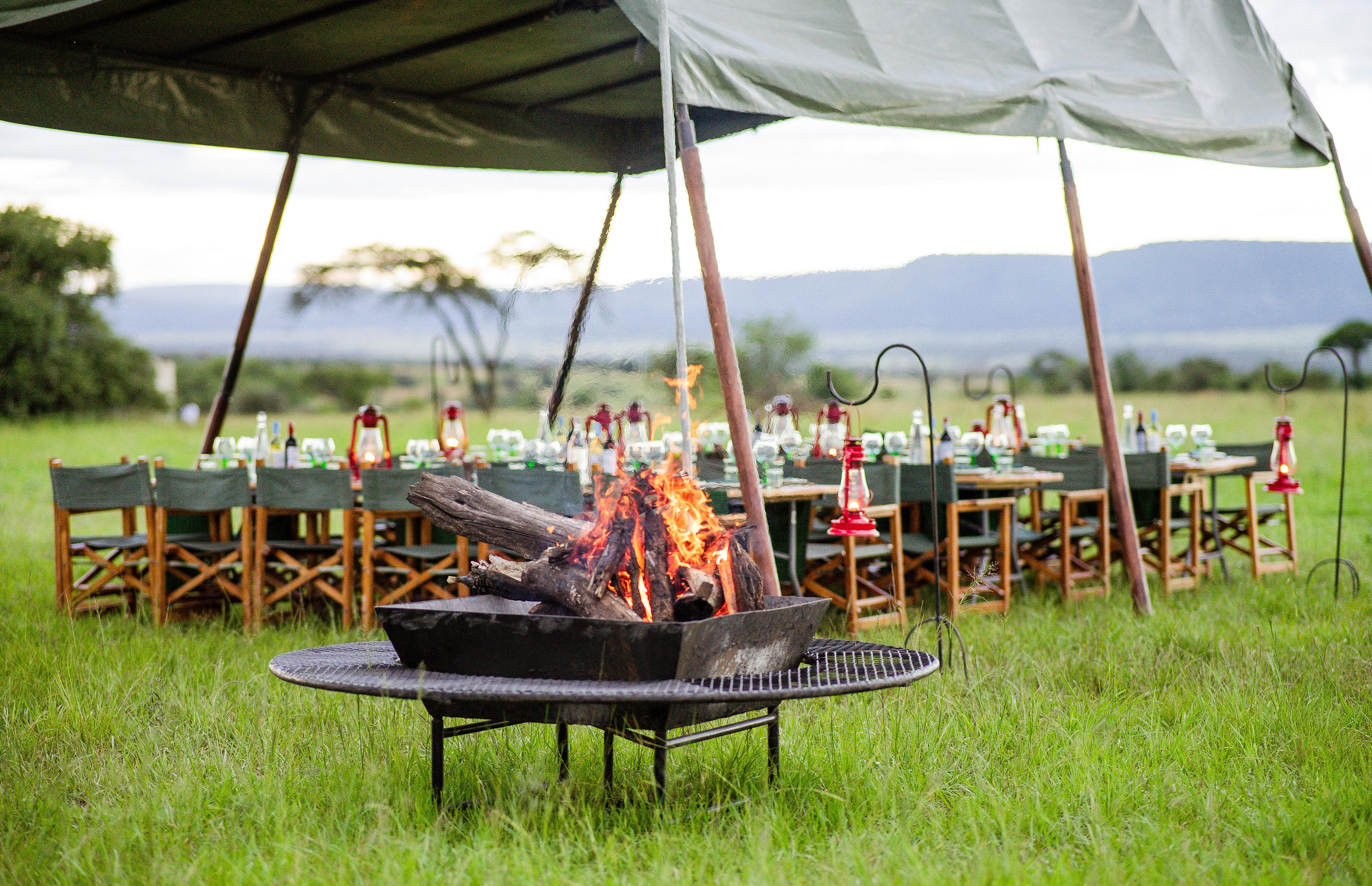 Glamping safari dinner with roaring campfire under a tent in a grassy field.