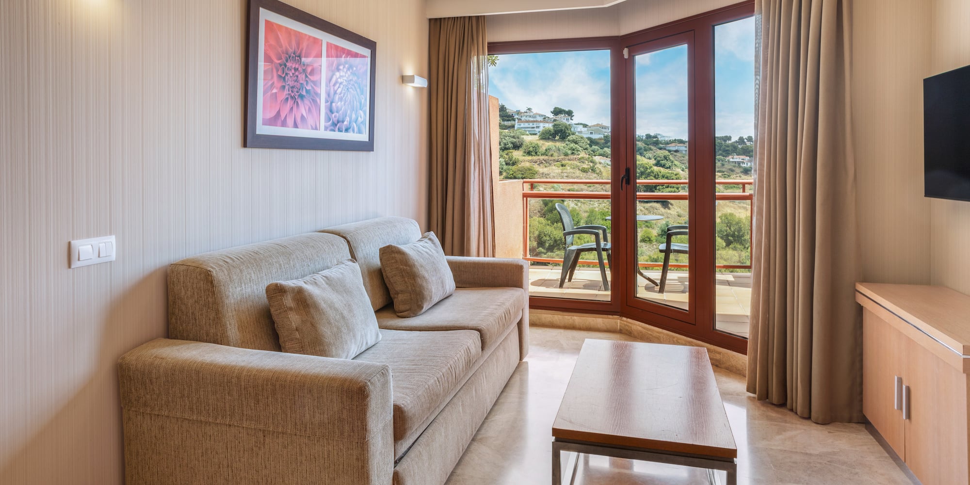 Modern living room with beige sofa, coffee table, and balcony overlooking hills.
