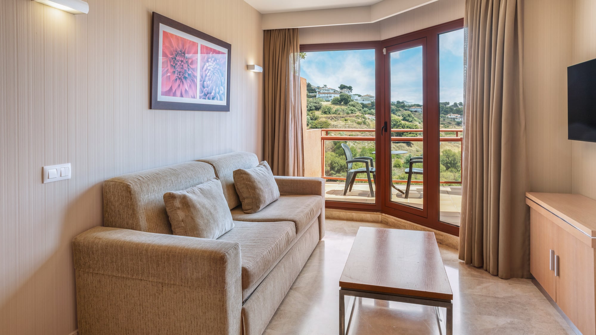 Modern living room with beige sofa, coffee table, and balcony overlooking hills.