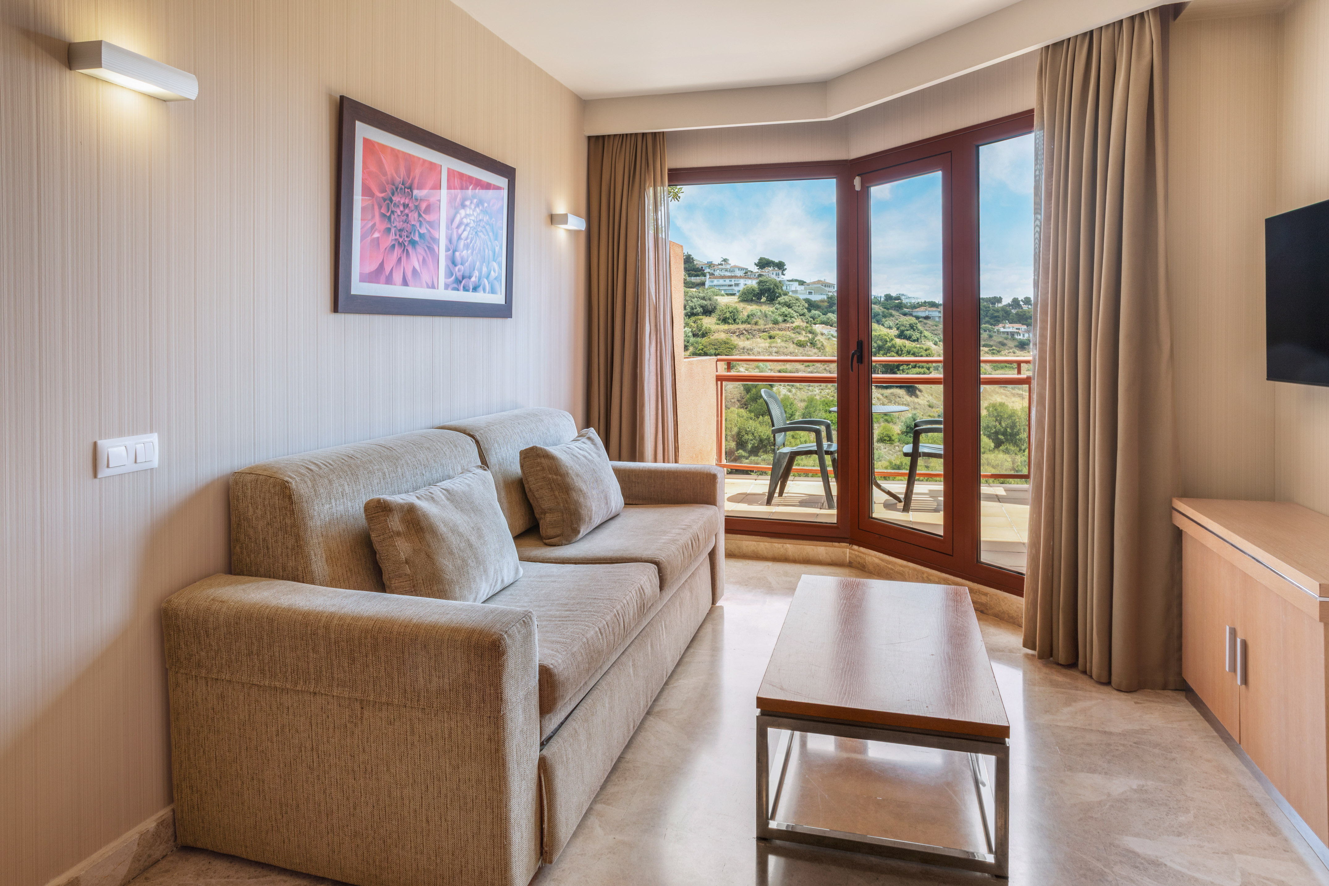 Hotel suite living room with sofa, balcony, and panoramic hill views.