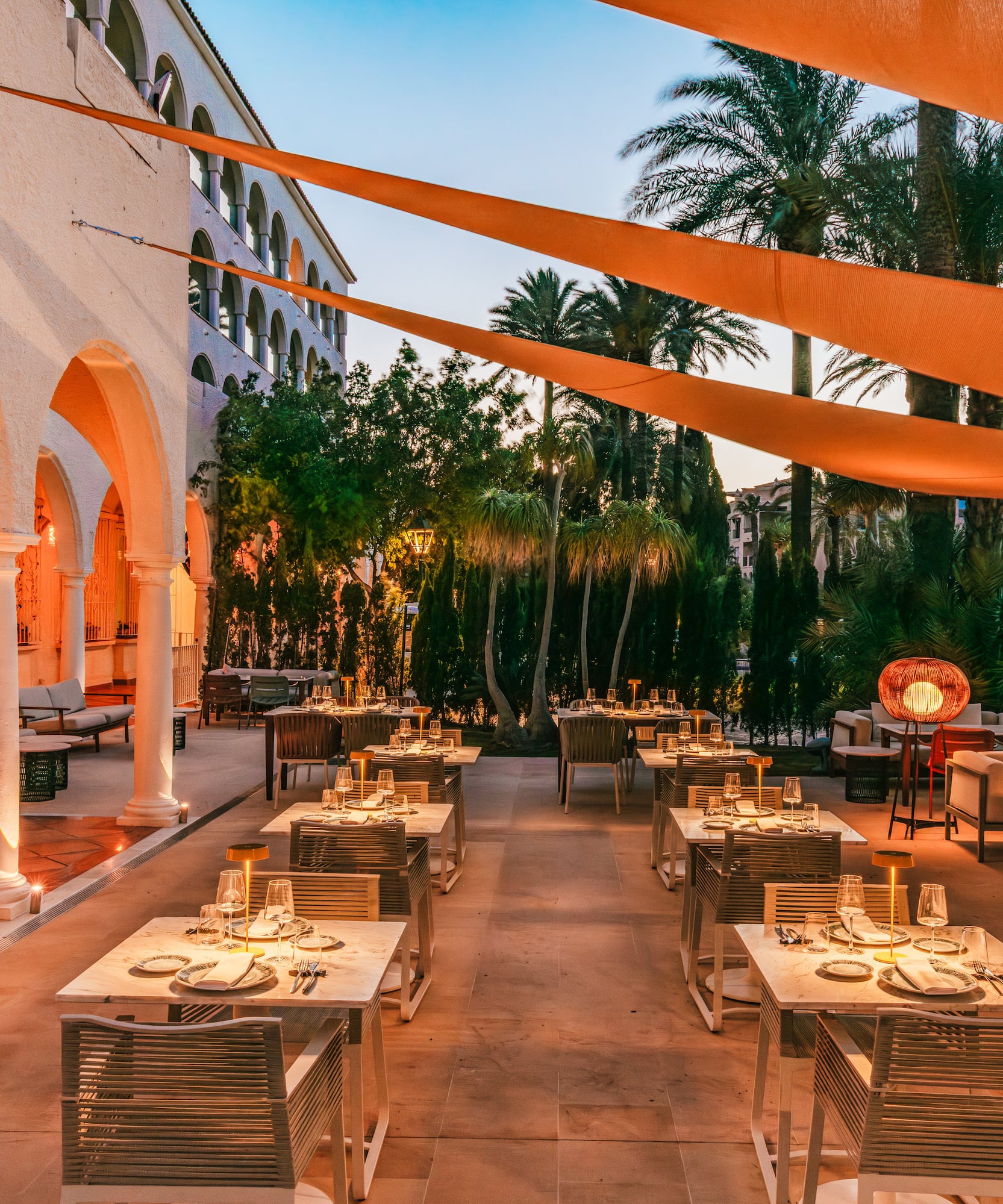Outdoor dining patio with set tables, palm trees, and warm evening lighting.