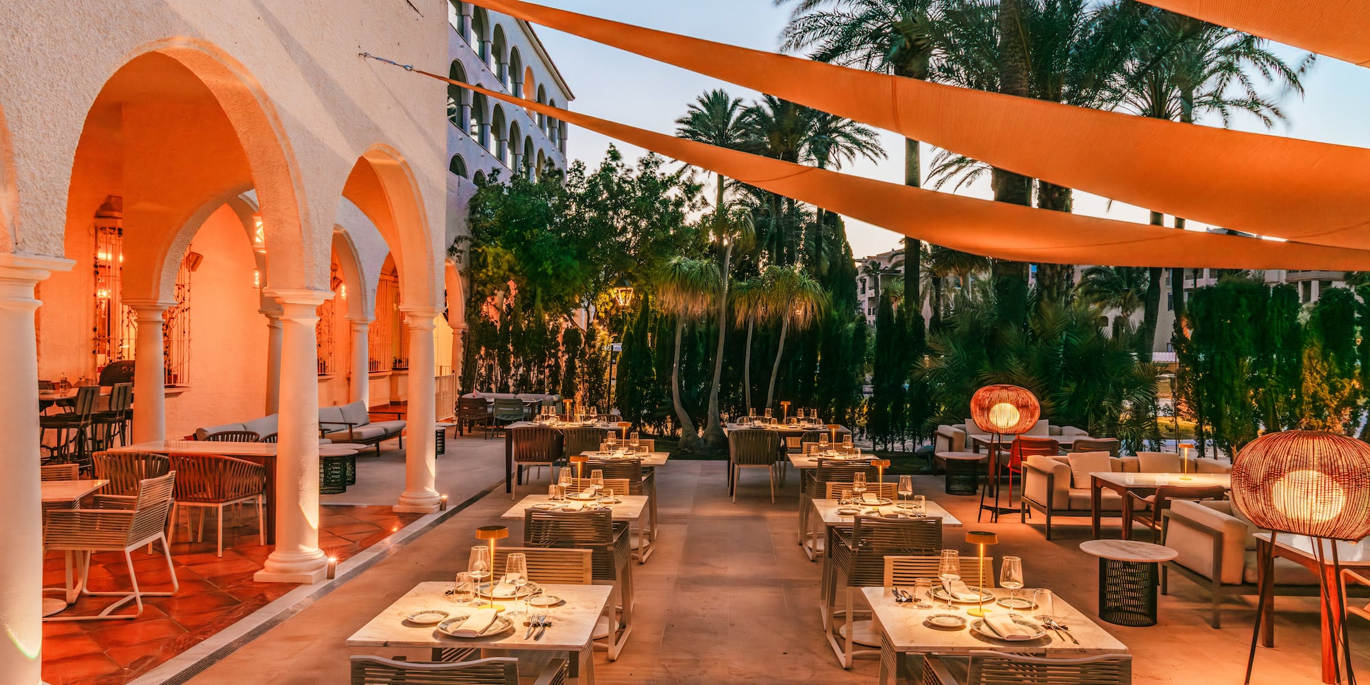 Outdoor dining patio with set tables, palm trees, and warm evening lighting.