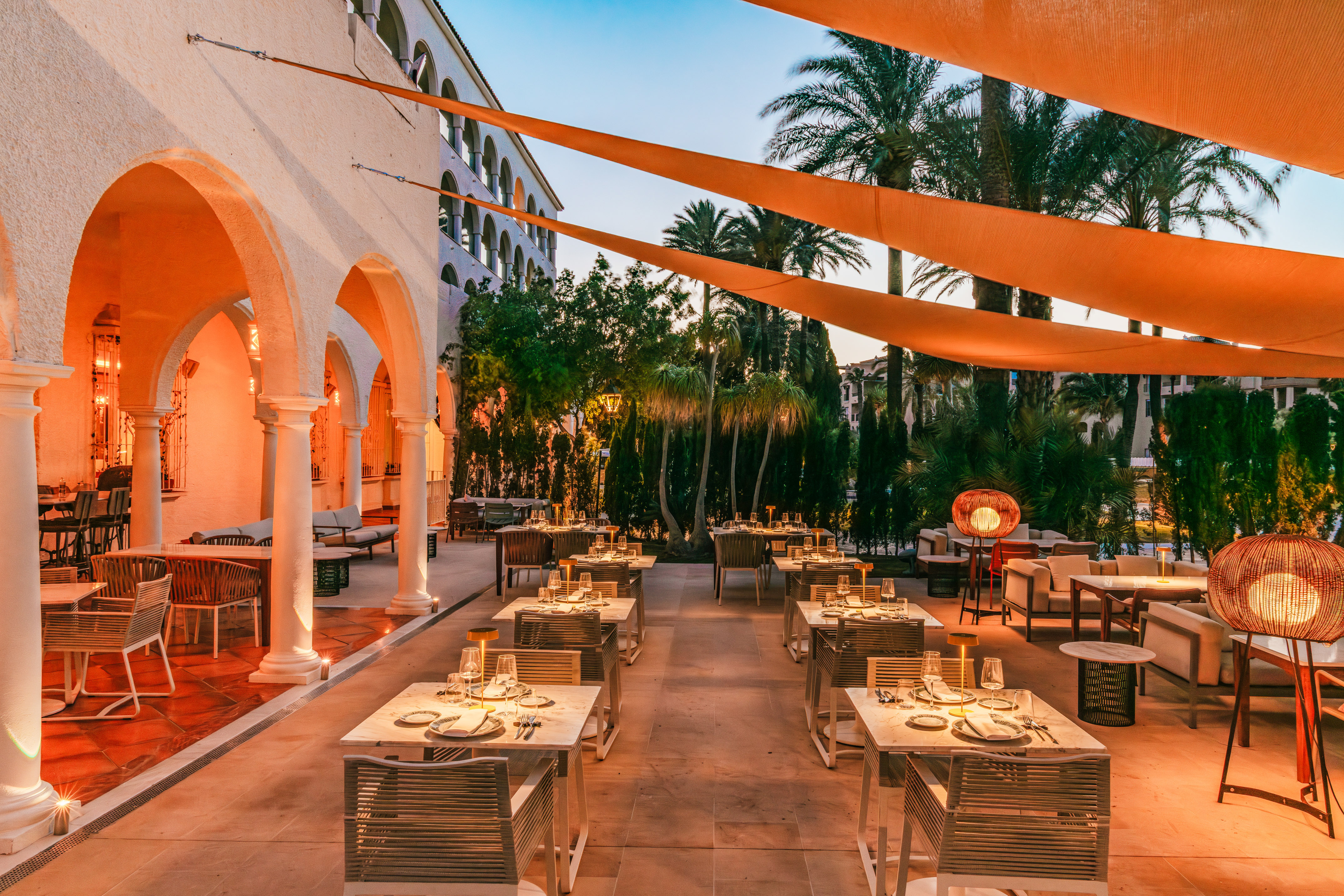 Outdoor dining patio with set tables, palm trees, and warm evening lighting.
