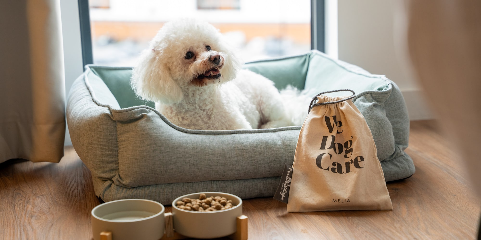 White fluffy dog in bed with Meliá pet amenities: food, water, and care kit.