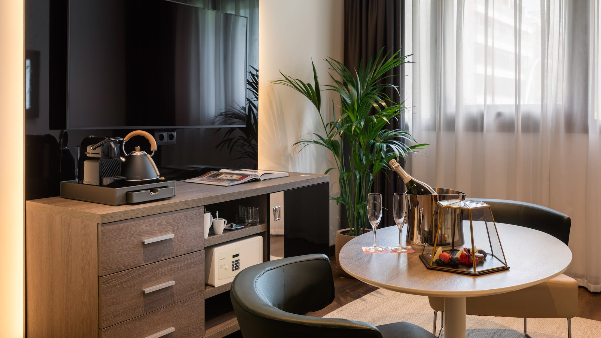 Habitación de hotel moderna con TV, cafetera, champagne, mesa y minibar.