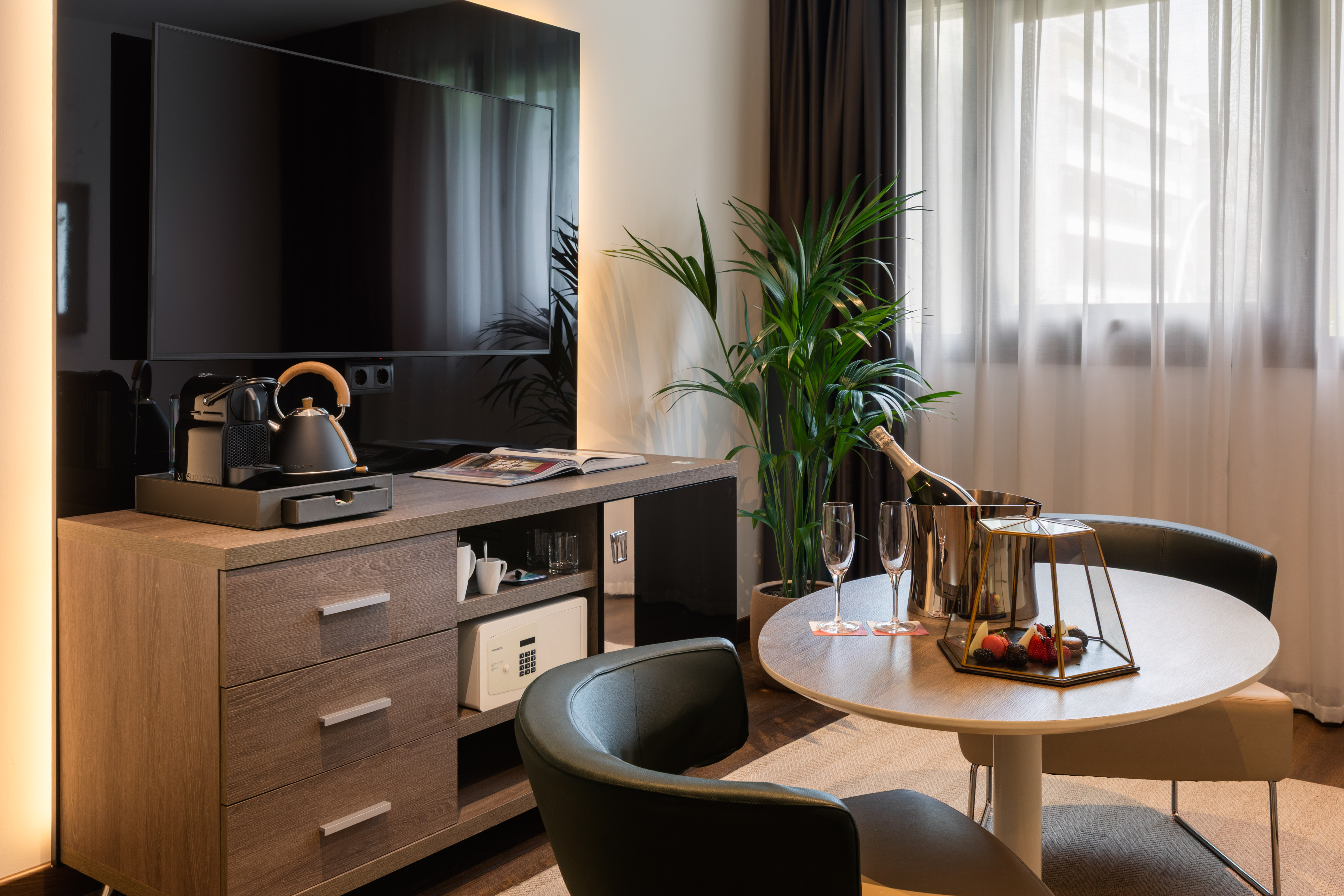Habitación de hotel moderna con TV, cafetera, champagne, mesa y minibar.