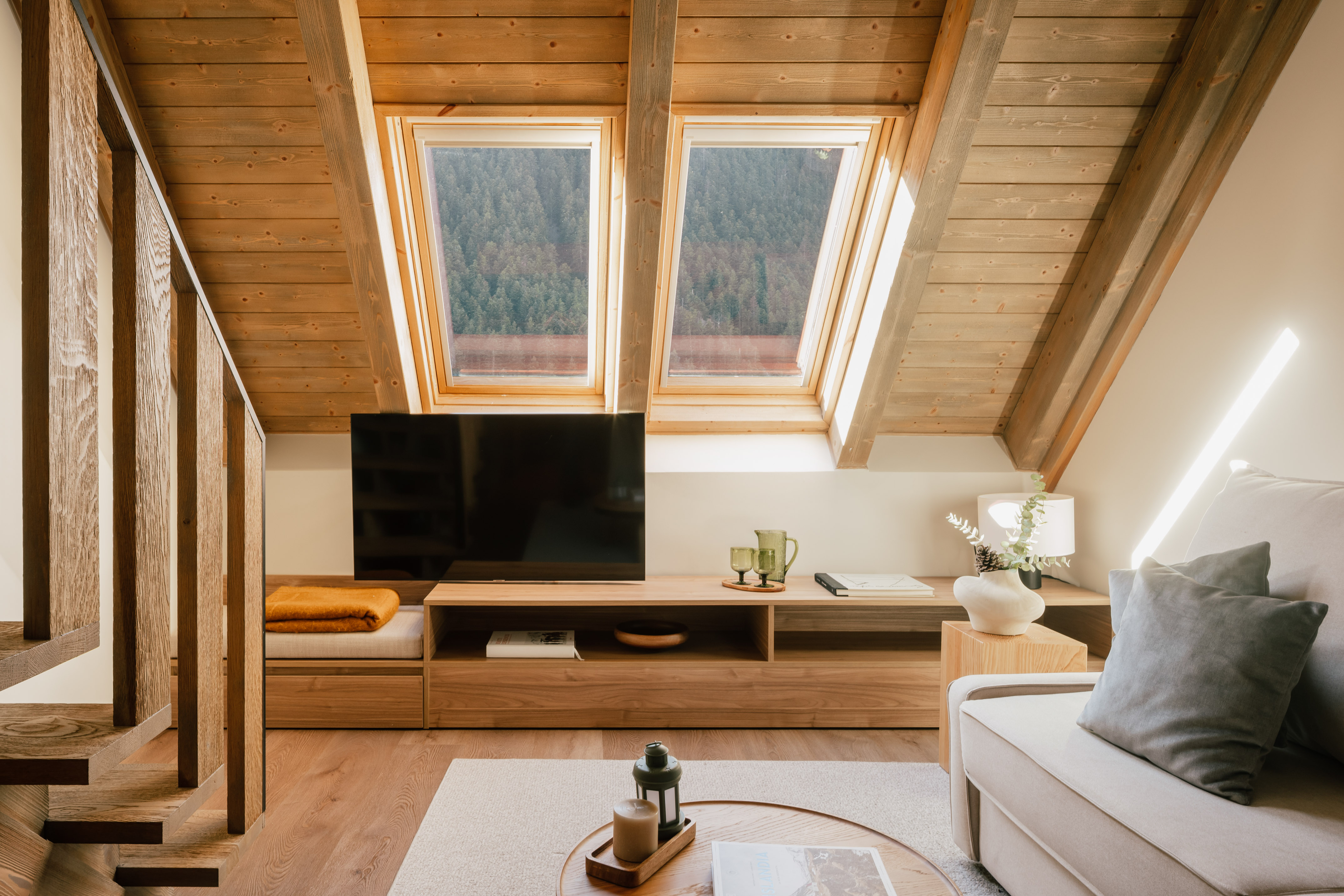 Cozy attic living room with wooden ceiling, skylights, forest view, TV, and sofa.