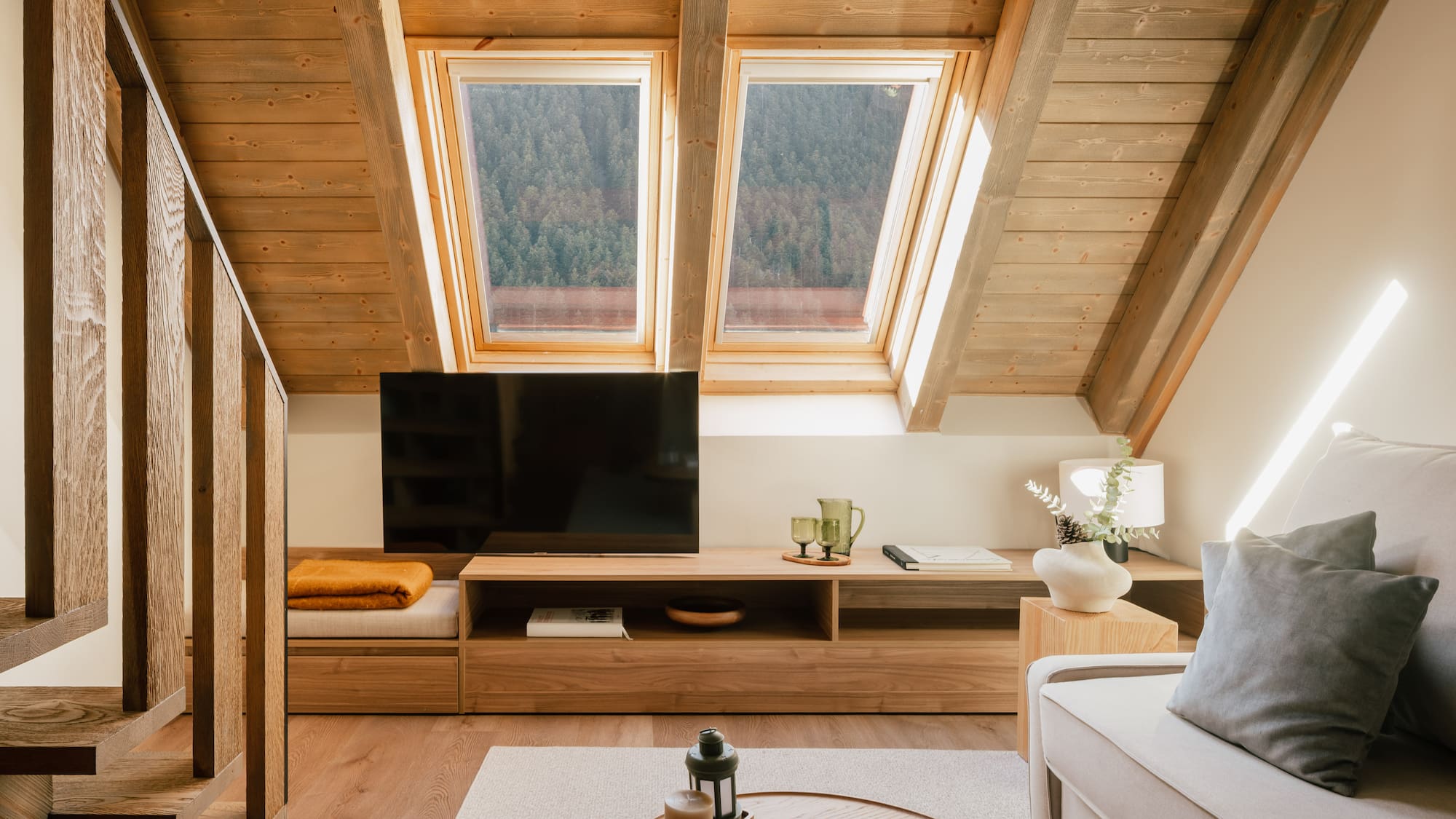 Cozy attic living room with wooden ceiling, skylights, forest view, TV, and sofa.