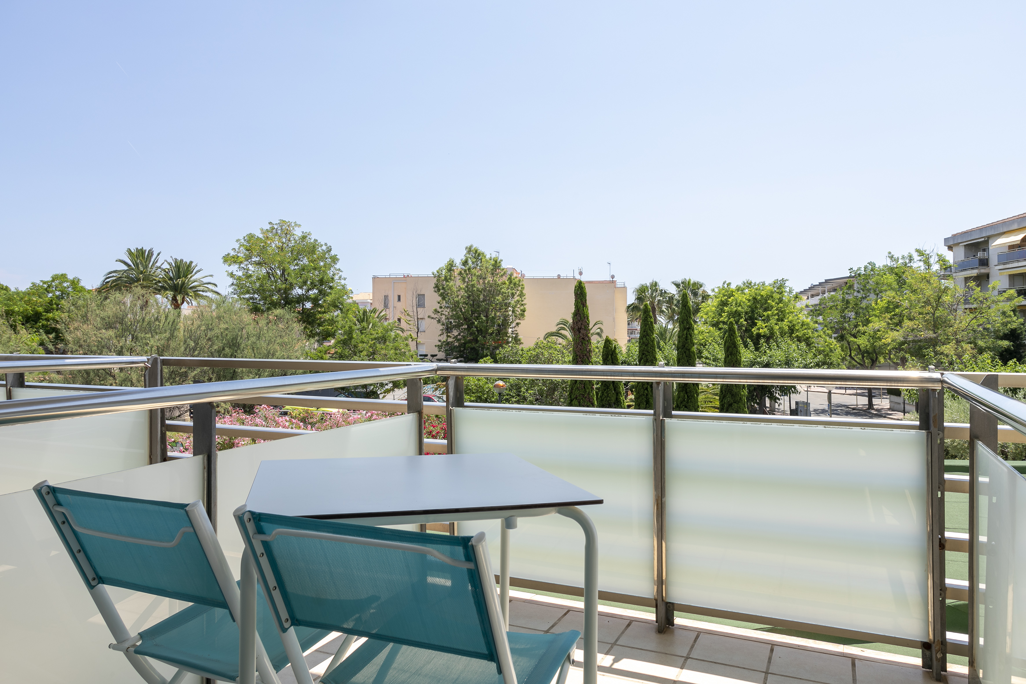 Balcony with turquoise chairs, table, sunny view of green trees and buildings.