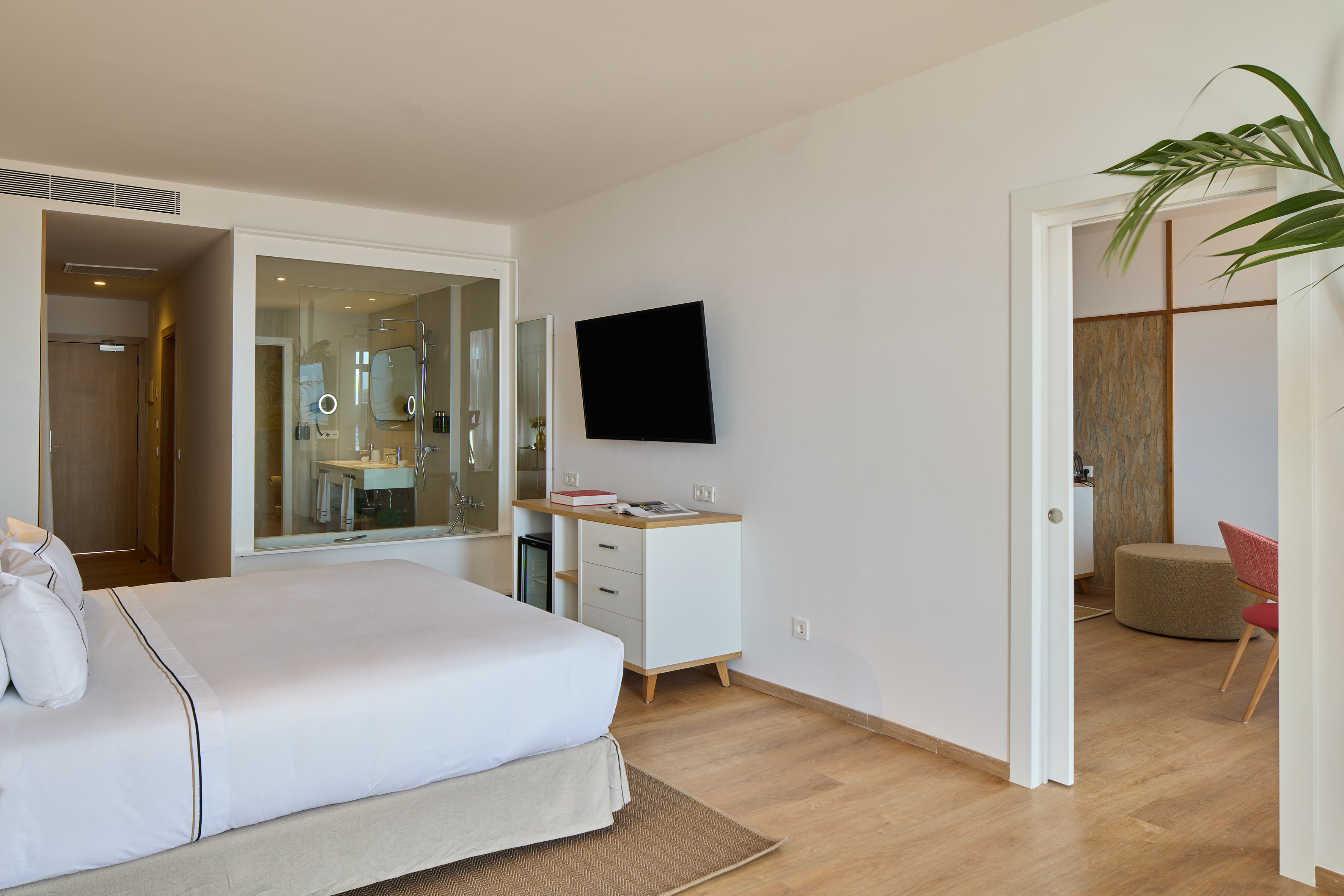 Modern hotel room with a white bed, wall-mounted TV, and glass bathroom wall.