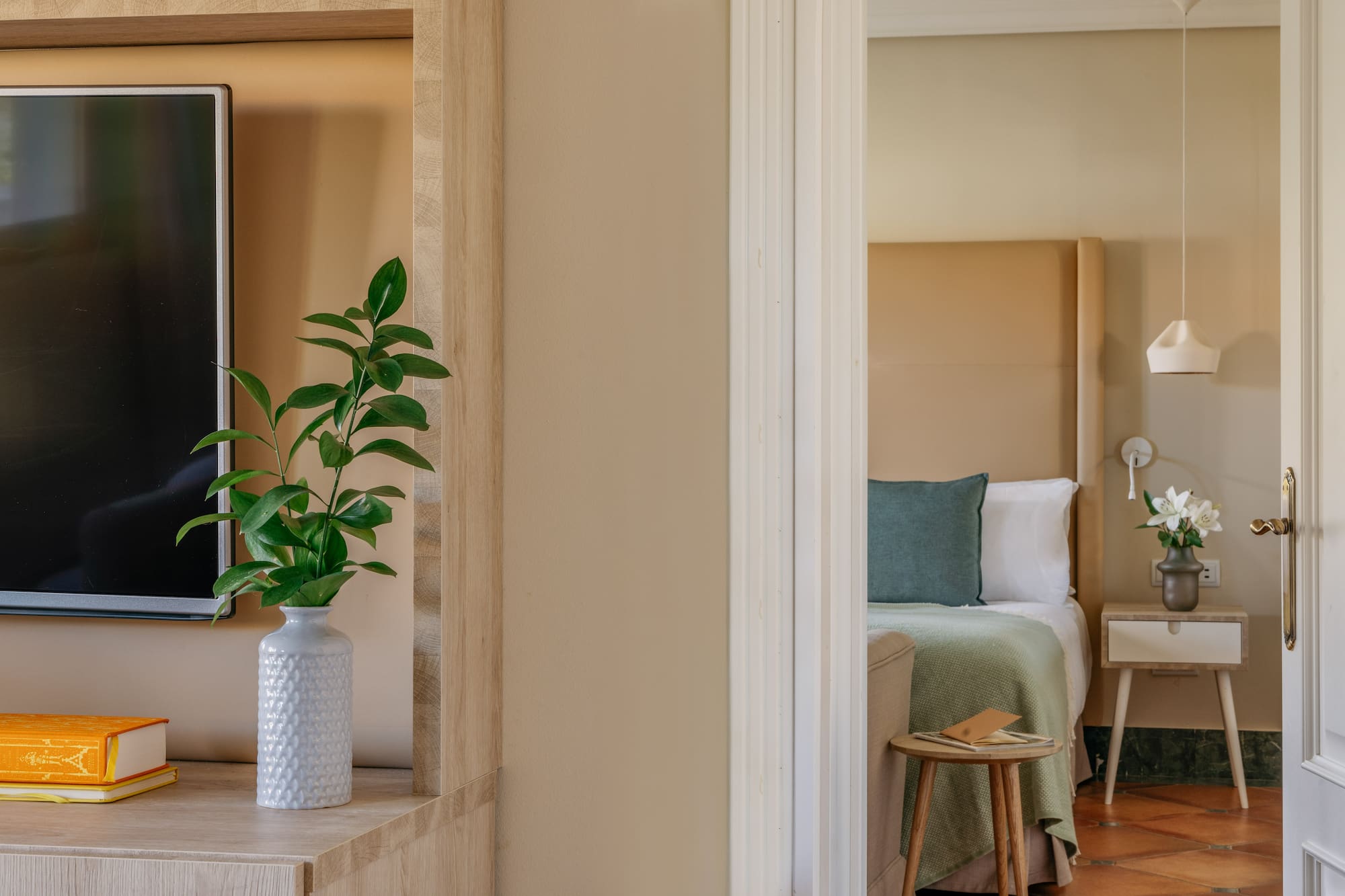 Modern hotel interior: plant, TV, and bedroom with bed and green blanket.