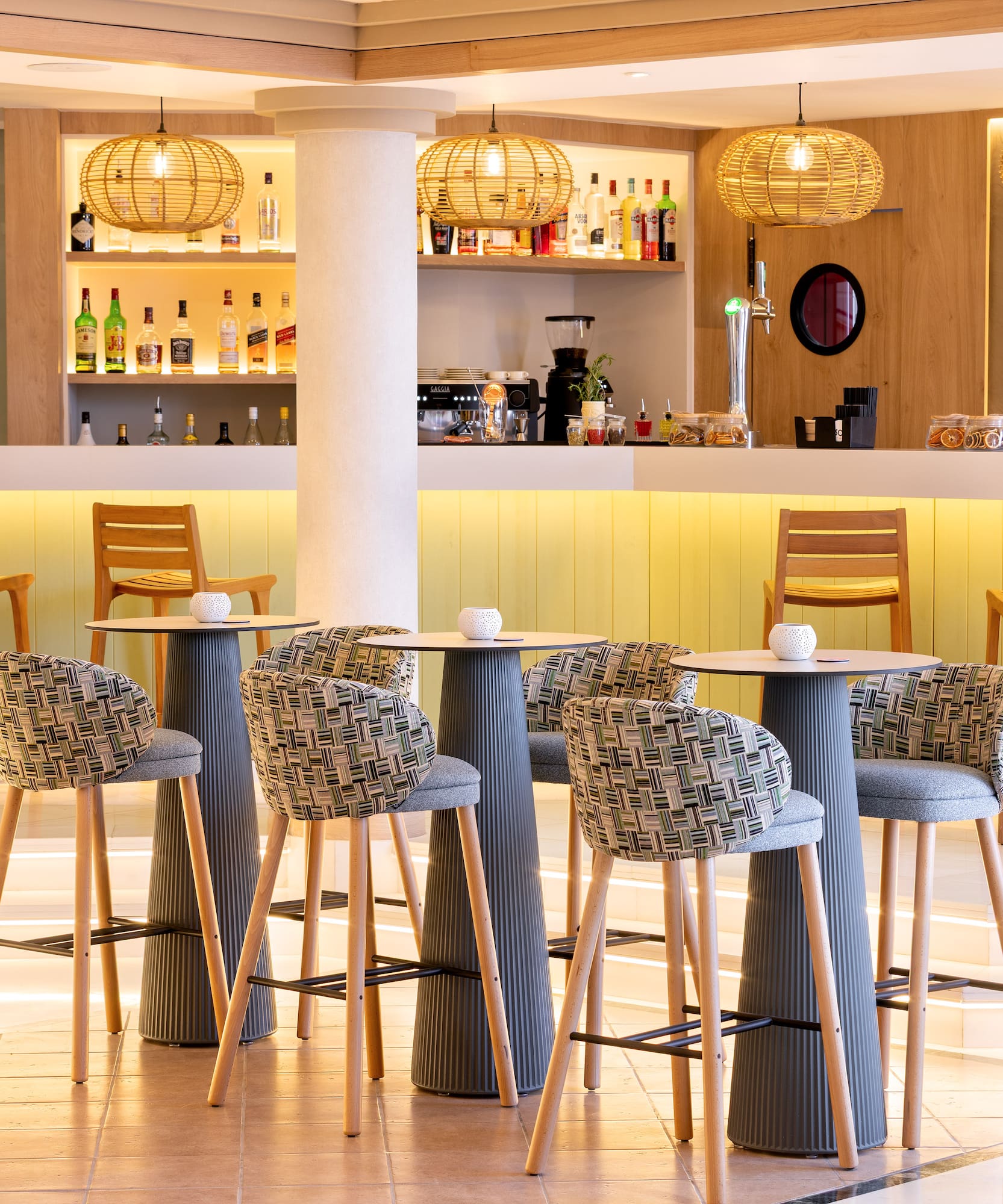Modern hotel bar with high tables, patterned chairs, and rattan pendant lights.