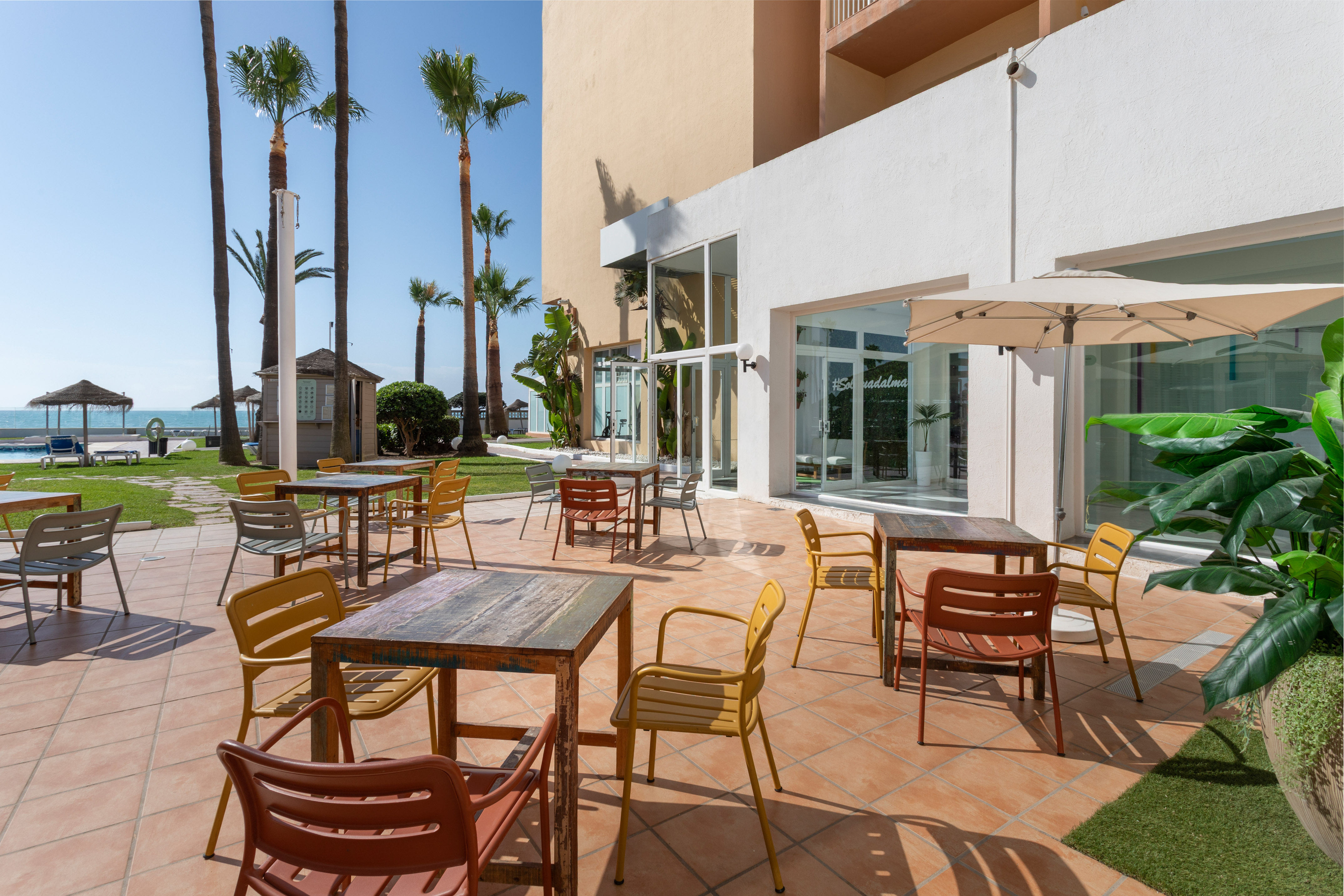 Terraza de hotel con mesas, sillas coloridas, palmeras y vistas al mar.