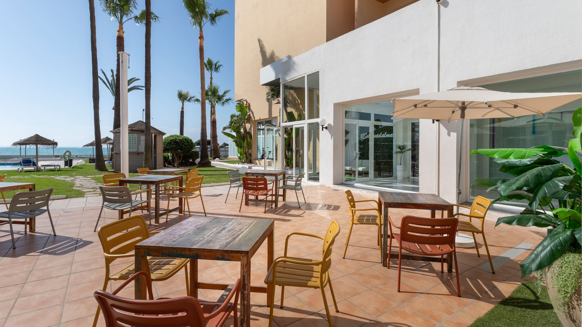 Terraza de hotel con mesas, sillas coloridas, palmeras y vistas al mar.