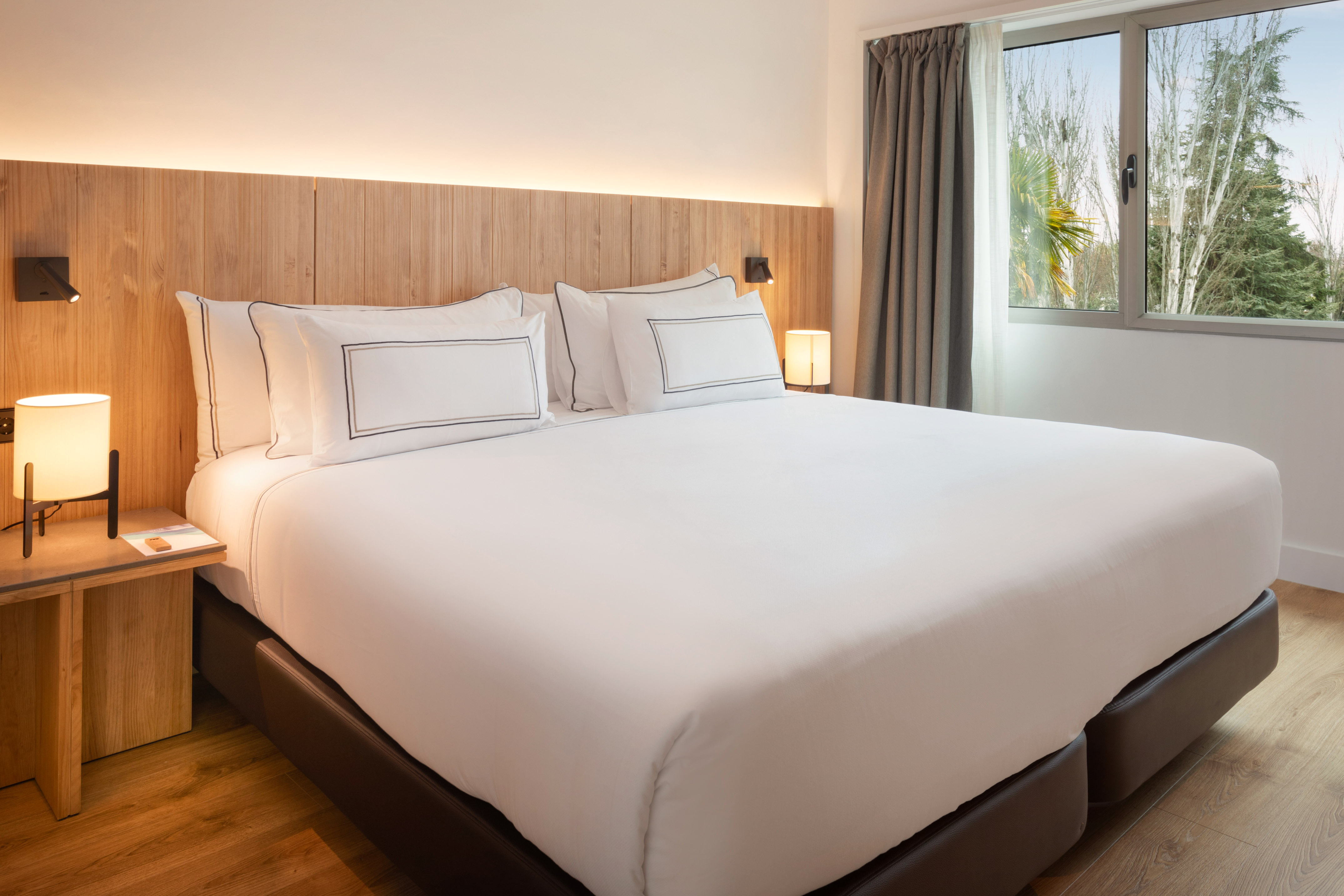 Modern hotel bedroom with king bed, wooden headboard, and natural light.
