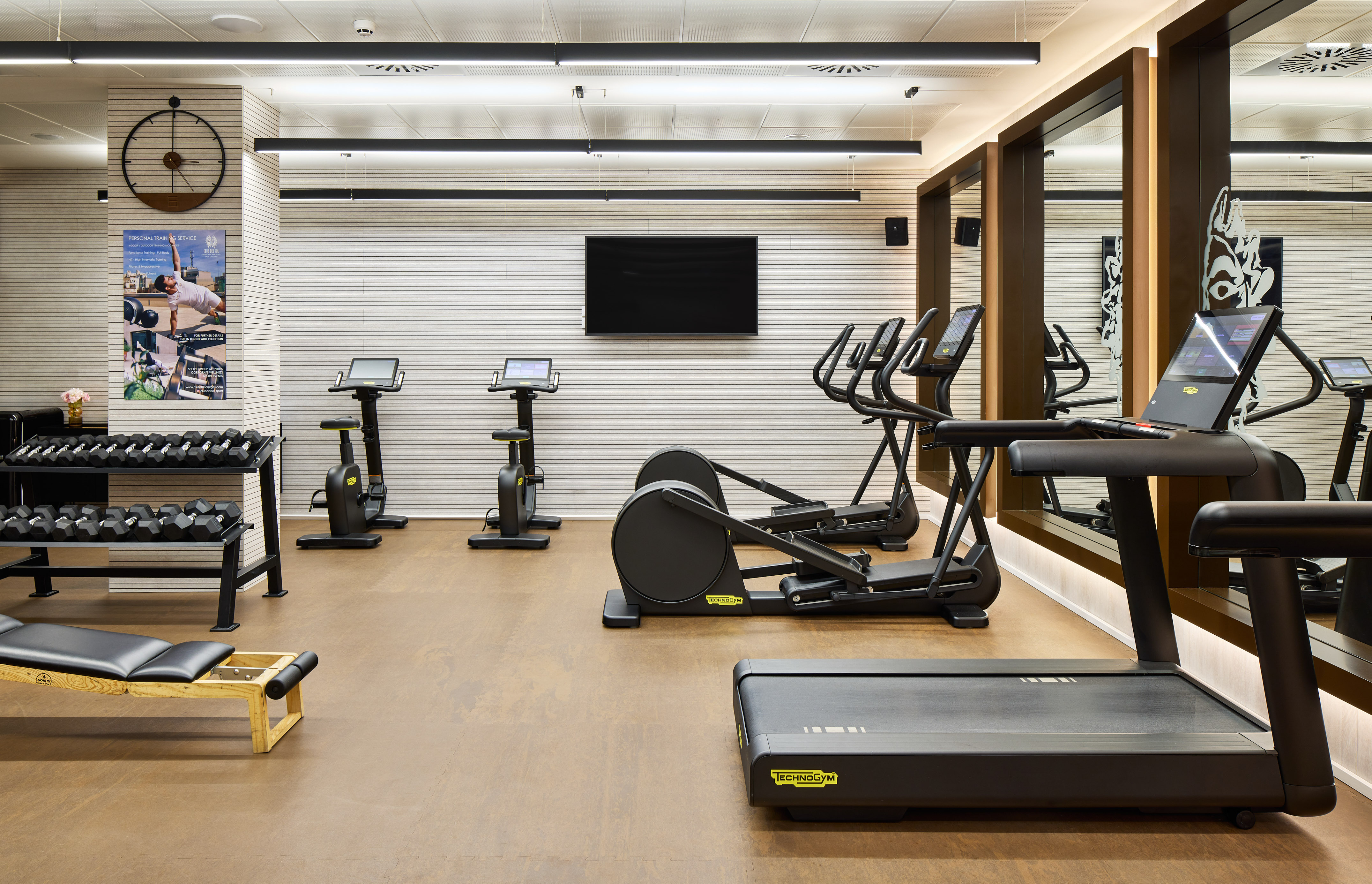 Modern gym with Technogym treadmill, ellipticals, bikes, and dumbbell rack.